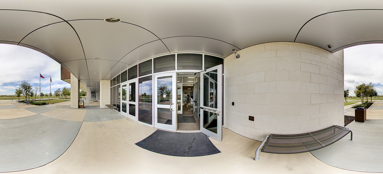 Panoramic view of a modern building entrance with glass double doors propped open, a covered walkway with benches, and flags visible outside along a landscaped campus pathway.