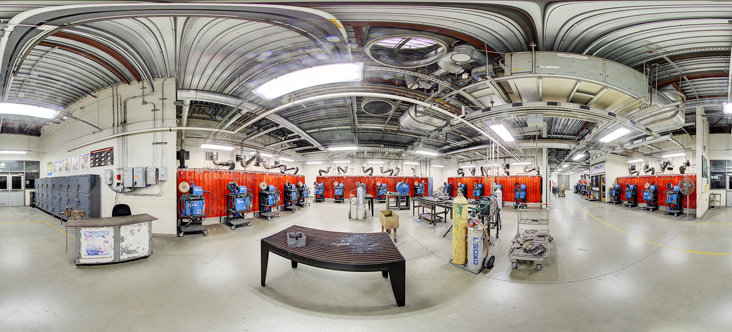 Panoramic view of a welding training lab with multiple blue welding stations lined along red safety curtains, ventilation ducts overhead, gas cylinders, and metal worktables arranged across an open industrial workspace.