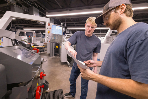 Two students stand beside a CNC machine, reviewing a sheet of paper together while discussing machining details in a workshop.
