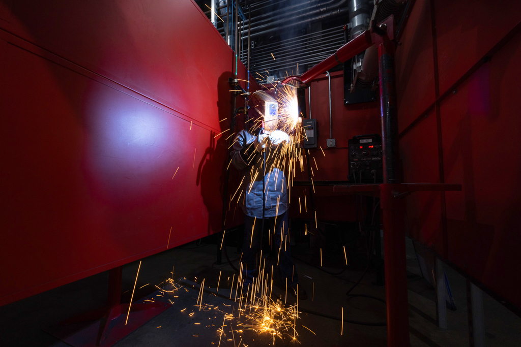 A student wearing a welding helmet works inside a red welding booth, creating a bright arc and a shower of sparks that scatter across the floor, with ventilation ducts and equipment surrounding the workspace.