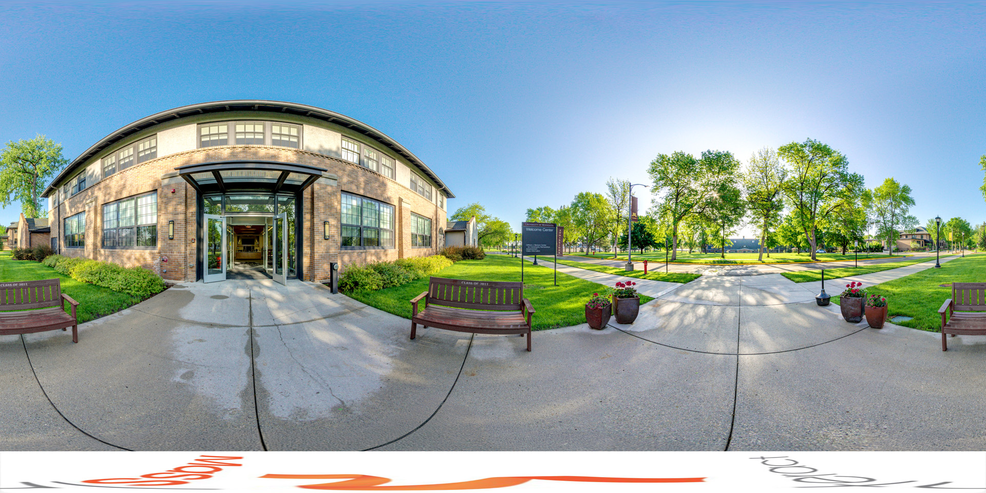 Panoramic view of a campus welcome center with a brick building, open glass doors, two benches, and a green lawn under a clear, sunny sky.