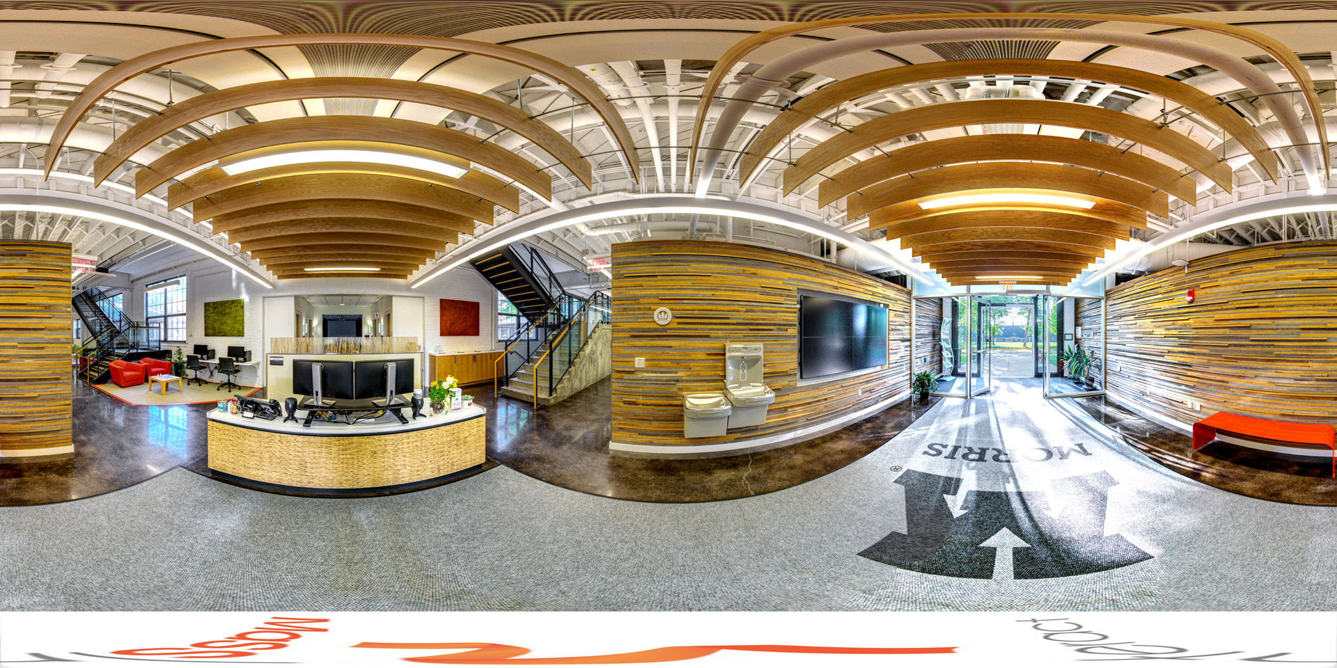 Panoramic view of the interior of a modern admissions office with a wood-paneled wall, curved ceiling beams, a reception desk, and a glass entrance door.