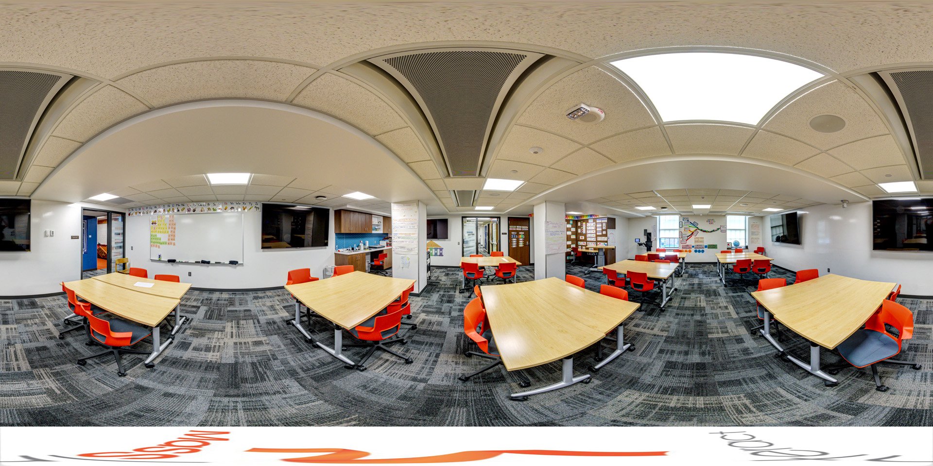 Panoramic view of a modern classroom with several wooden tables, bright orange chairs, and large display screens on the walls. The room has a whiteboard, bookshelves, and fluorescent ceiling lights.