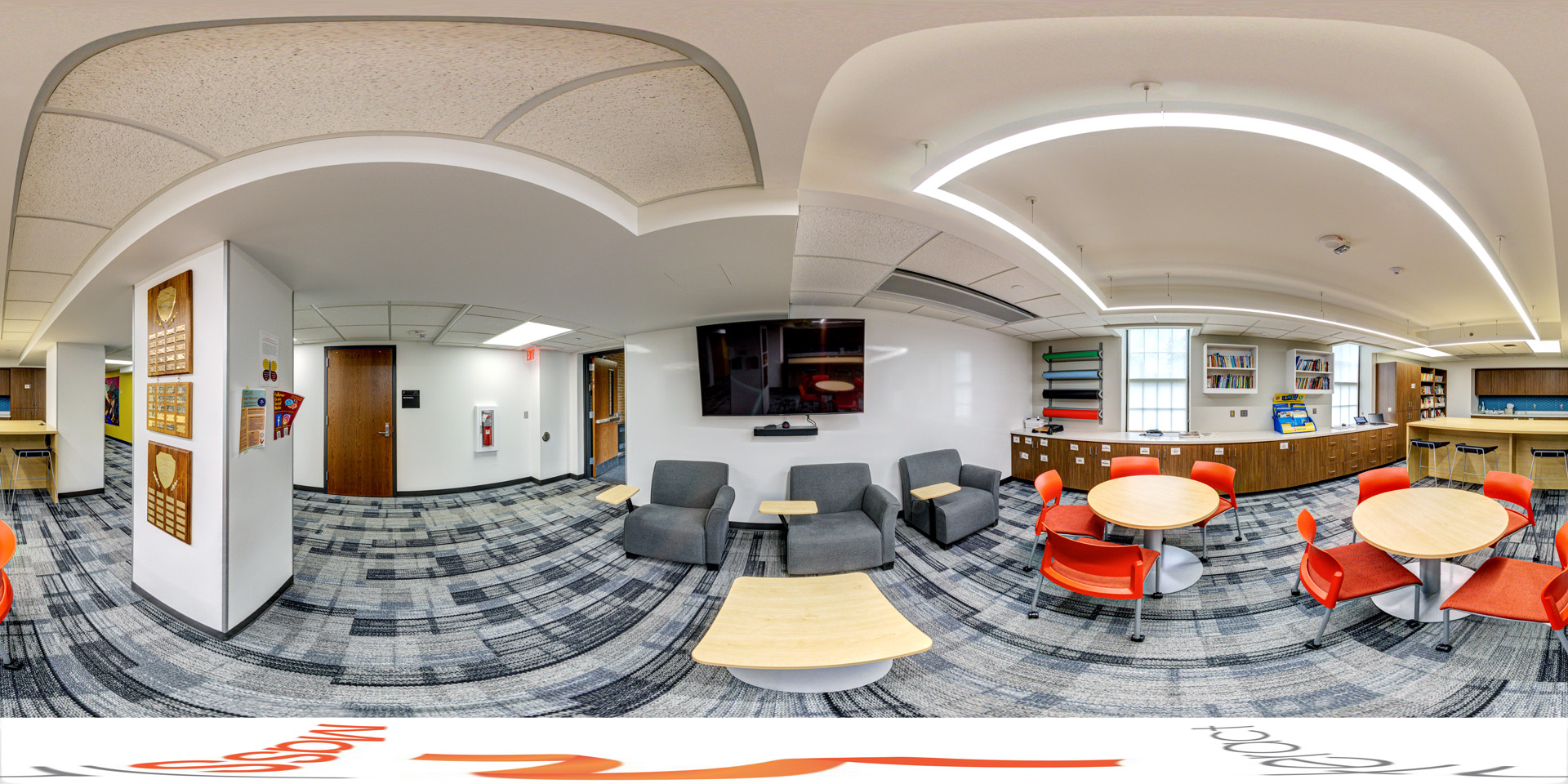 Panoramic view of a collaborative lounge with round tables, bright orange chairs, and a seating area with gray armchairs. The room features a wall-mounted TV, bookshelves, and a counter with stools along the back wall.
