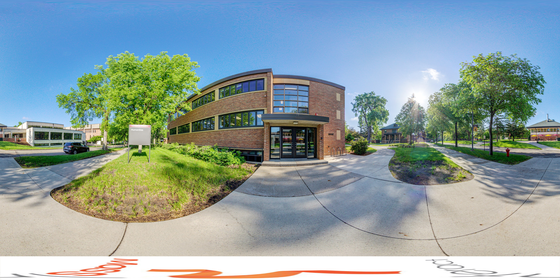 Panoramic view of an outdoor view of a campus building labeled 'Humanities,' with a brick exterior, large windows, and a modern design. The surrounding area features trees, pathways, and a bright, sunny sky.