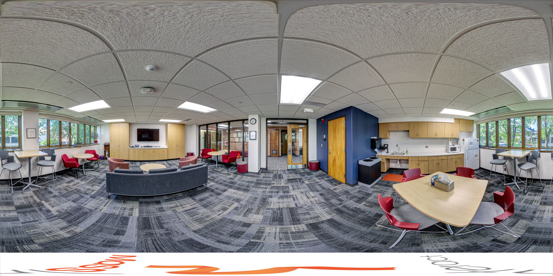 Panoramic view of a lounge area with modern furniture, including gray and red chairs, round tables, and a kitchenette with a microwave and sink. The room is well-lit by large windows and overhead lights, and a TV is mounted on the wall.