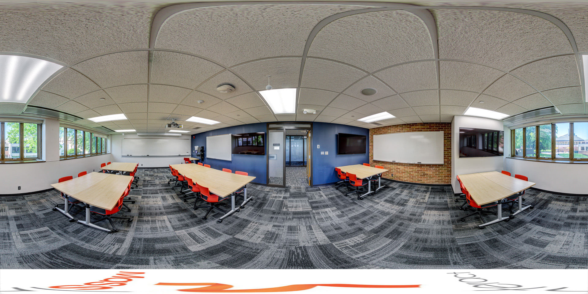 Panoramic view of a classroom with large windows, rectangular tables, bright orange chairs, and whiteboards on the walls. The room also features a mounted display screen and overhead projector, with a modern carpeted floor and ceiling lights.