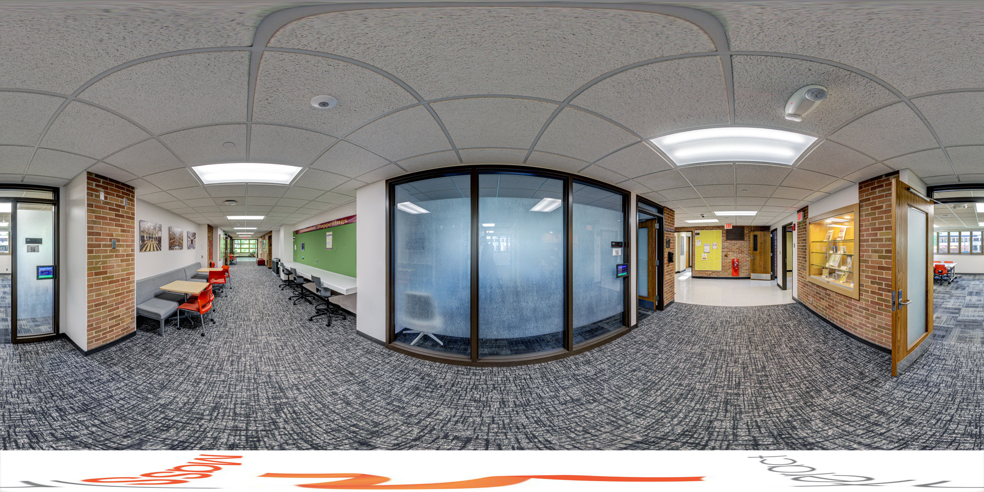 Panoramic view of a hallway with frosted glass meeting rooms on the right, desks with chairs along the left, and soft seating areas with tables. The corridor is carpeted and decorated with framed photos, with brick walls and bright overhead lighting.