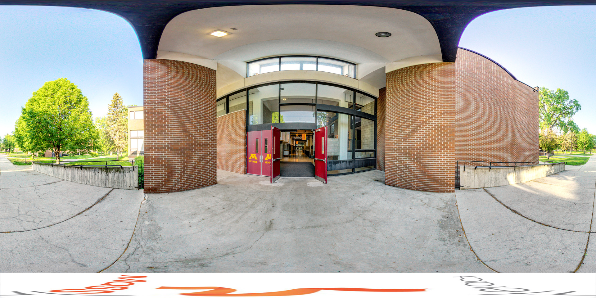 Panoramic view of an exterior view of the Humanities Fine Arts building entrance, featuring red double doors with the University of Minnesota Morris logo. The building has large windows and brick walls, and the surrounding area includes a tree-lined walkway under a clear, sunny sky 