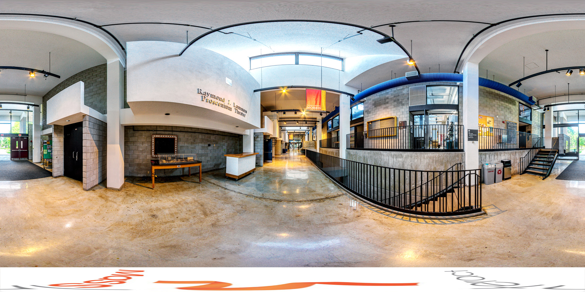 Panoramic view of the interior of the Humanities Fine Arts building, featuring a polished floor, curved walls, and multiple levels with staircases. The entrance to the 'Raymond J. Lammers Proscenium Theatre' is visible, along with a brightly lit hallway leading deeper into the building.