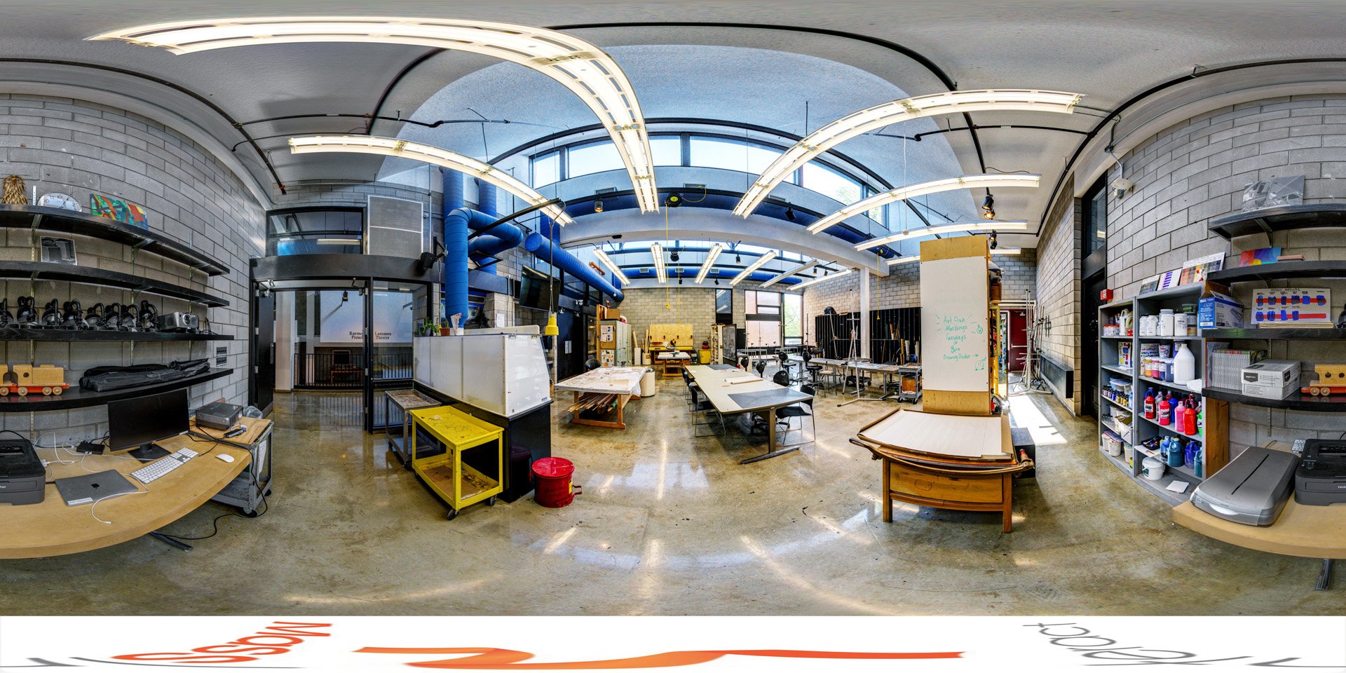 Panoramic view of an art studio with tables, shelves stocked with paint and supplies, and various art materials. The room has an industrial design with exposed pipes, high windows, and bright overhead lights. The workspace includes areas for painting and drawing, as well as computers and printers for digital work 