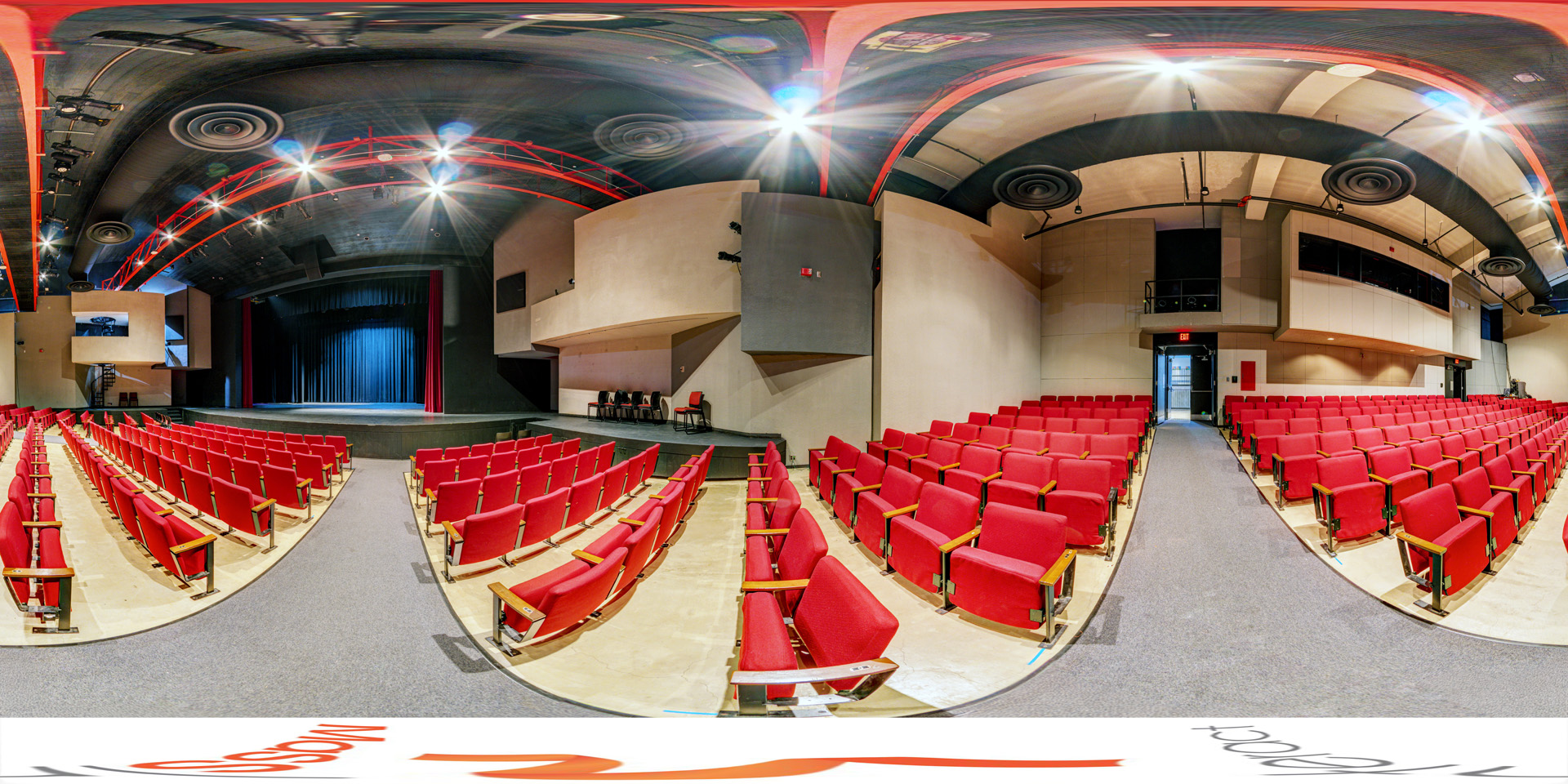 Panoramic view of a theater with rows of red seats facing a stage with closed blue curtains. The ceiling has exposed red beams and industrial lighting, and the walls feature a balcony and sound booth. The space is well-lit with a central aisle leading toward the stage. 
