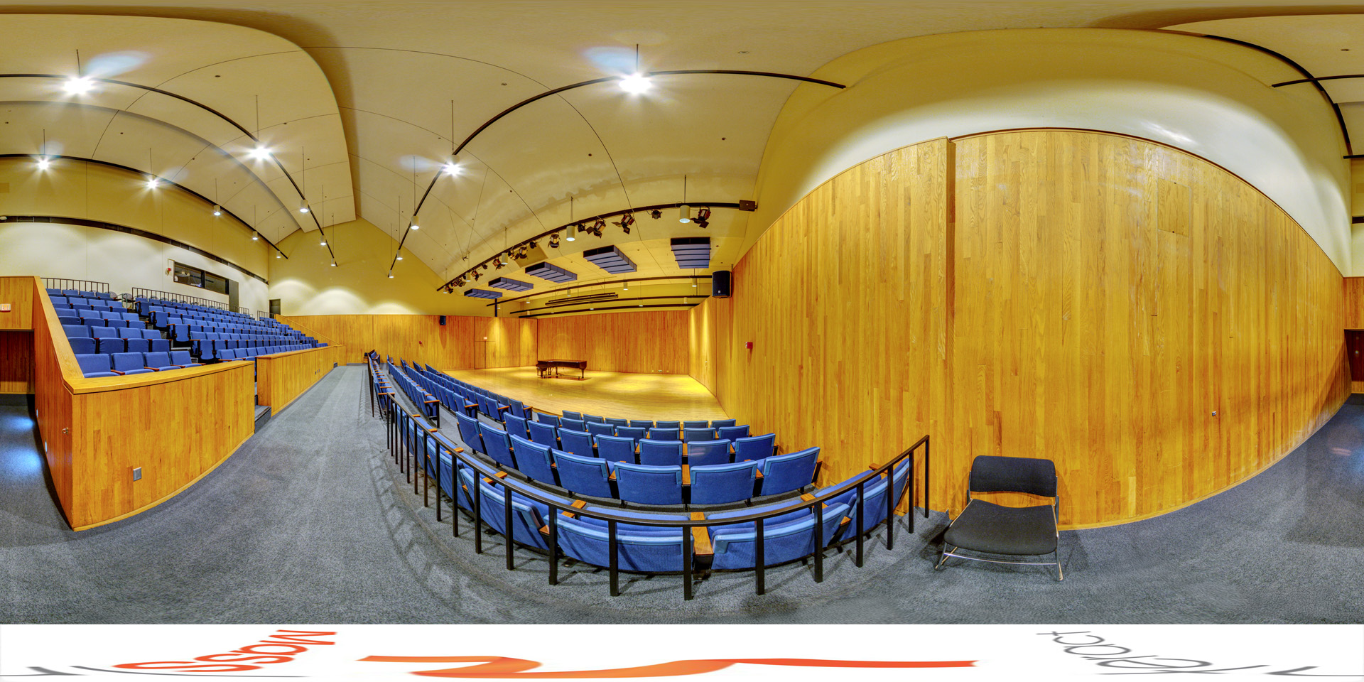 Panoramic view of a ecital hall with tiered seating featuring blue chairs and wooden paneling on the walls. The stage at the front has a grand piano, and the ceiling is equipped with lighting and acoustic panels. The room has a warm, intimate atmosphere designed for performances.