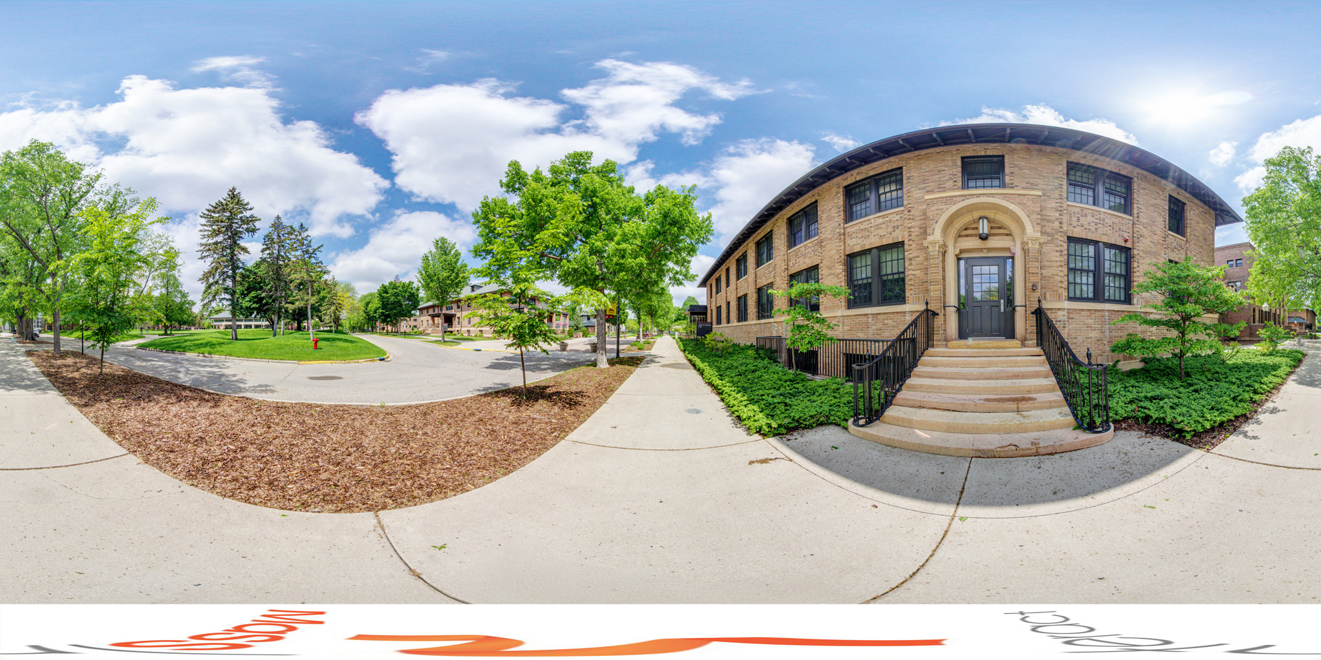 Panoramic view of an outdoor view of John Q. Imholte Hall, a brick building with arched entryway steps, surrounded by greenery. The scene includes trees, a sidewalk, and a bright, partly cloudy sky. 