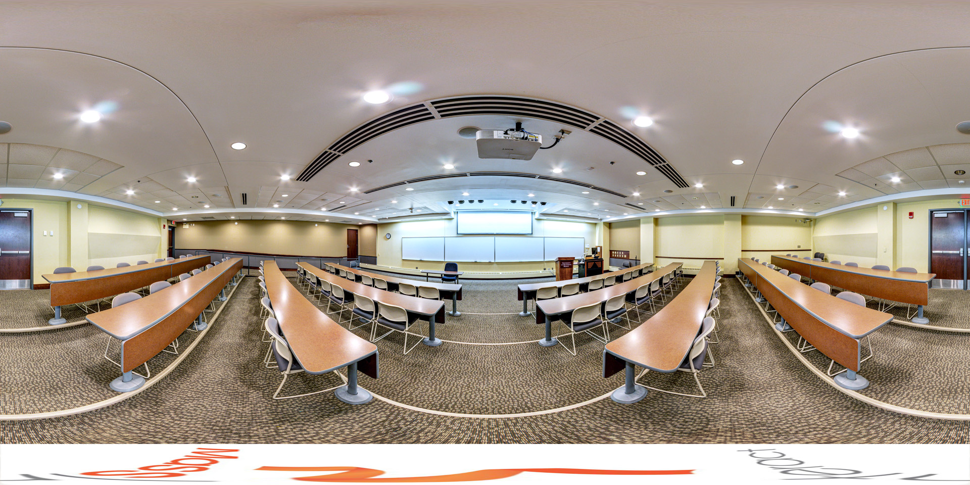 Panoramic view of a lecture hall with tiered seating arranged in curved rows, each equipped with long desks and chairs. The room features a projector and large whiteboards at the front, along with soft overhead lighting and a neutral color scheme.