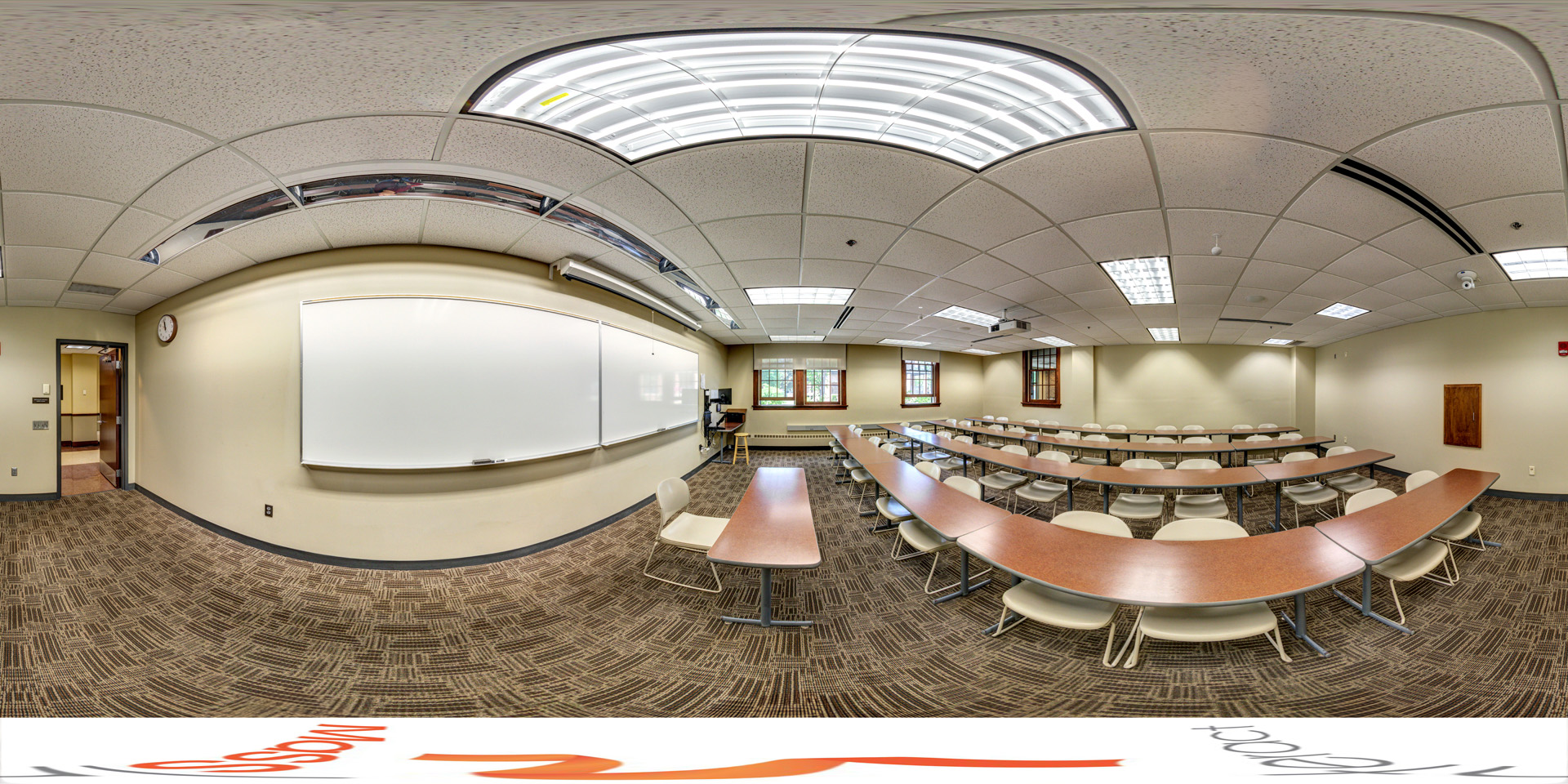 Panoramic view of a classroom with curved rows of desks and chairs facing a whiteboard at the front. The room is lit by fluorescent lights and features carpeted floors and windows that let in natural light. A projector is mounted on the ceiling for presentations.