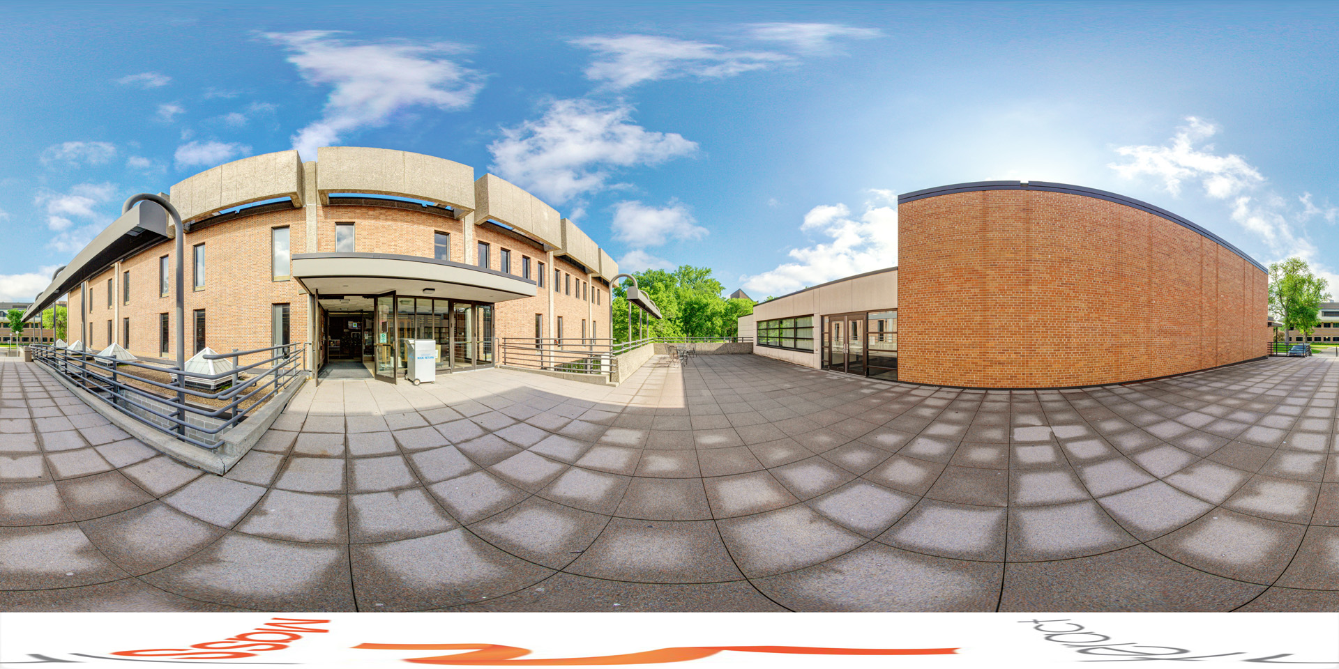 Panoramic view of an outdoor view of Rodney A. Briggs Library, featuring a brick exterior with large windows and a modern entryway. The surrounding area includes a spacious tiled patio under a partly cloudy sky.