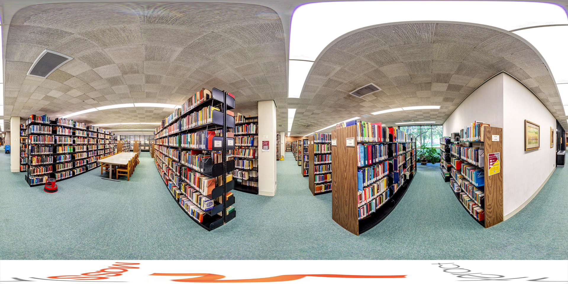 Panoramic view of a library section with tall bookshelves filled with books, organized in rows known as 'stacks.' The area has study tables with wooden chairs in the center, surrounded by shelves. The ceiling features a checkered pattern with fluorescent lighting, and the floor is carpeted. A large window in the background.