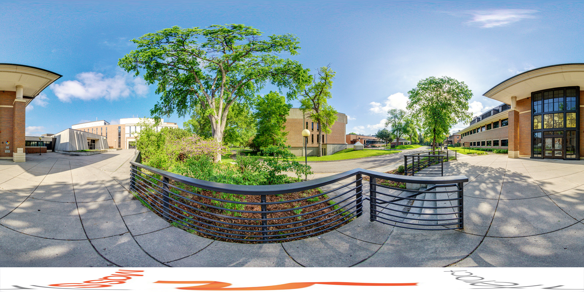 Panoramic view of an outdoor view of a campus area featuring modern academic buildings, including one with large windows and brick columns. The walkway is lined with trees, and there is a metal railing along the edge of the path. The sky is clear and sunny, casting shadows on the pavement. 