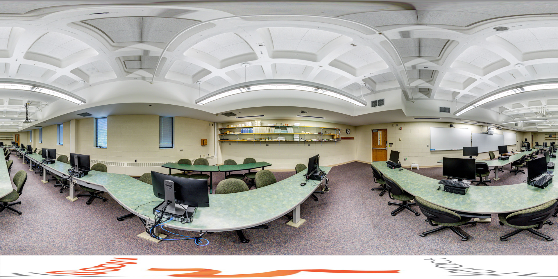 Panoramic view of a computer lab with curved green desks, desktop computers, and swivel chairs, featuring whiteboards and shelves along the walls.