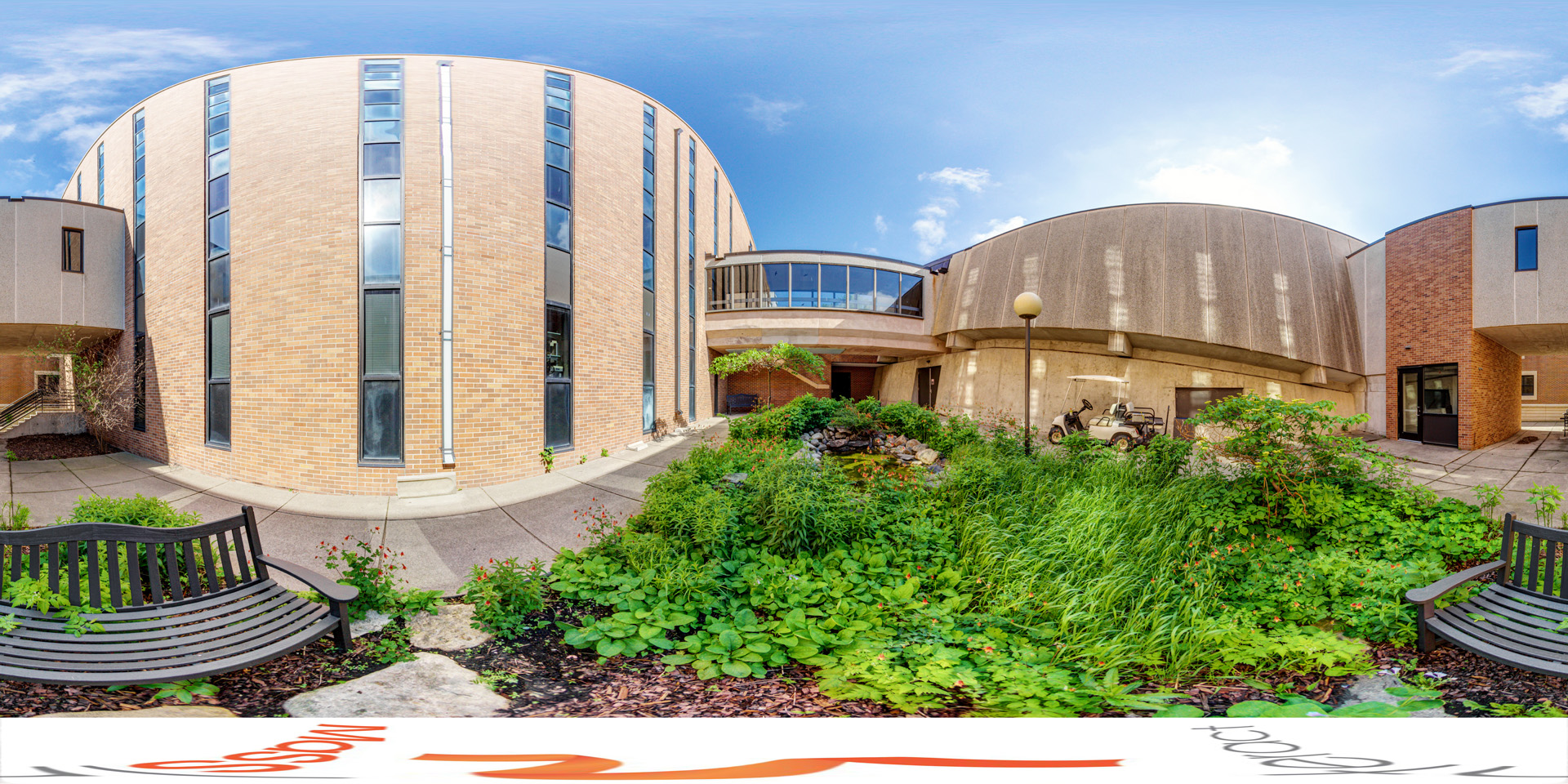 Panoramic view of a small garden with lush greenery and a pond, surrounded by brick and concrete buildings, featuring a black bench in the foreground.