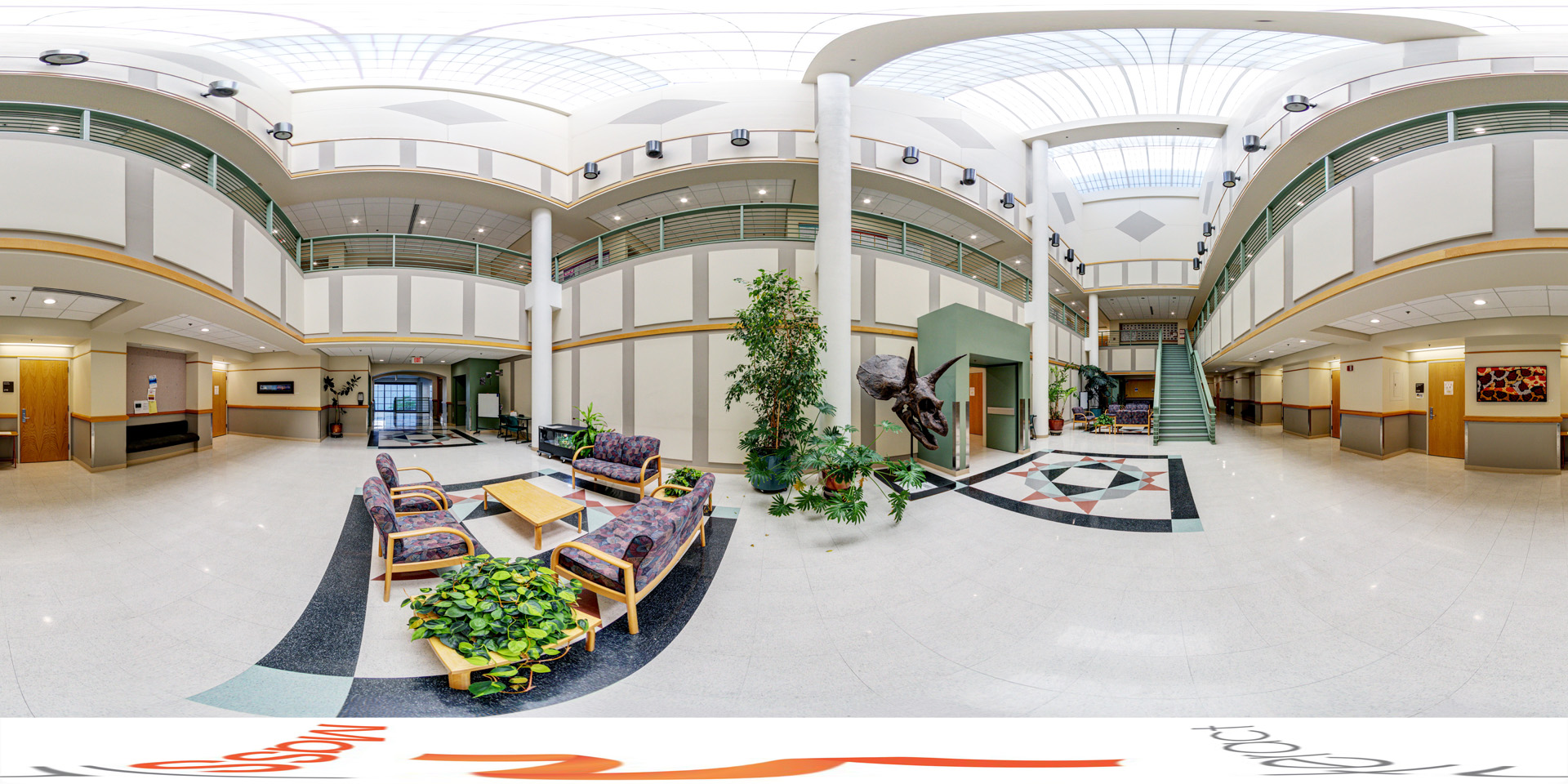 Panoramic view of a spacious atrium with multiple floors, lounge seating, a staircase, and a large dinosaur head sculpture hanging near a potted plant.