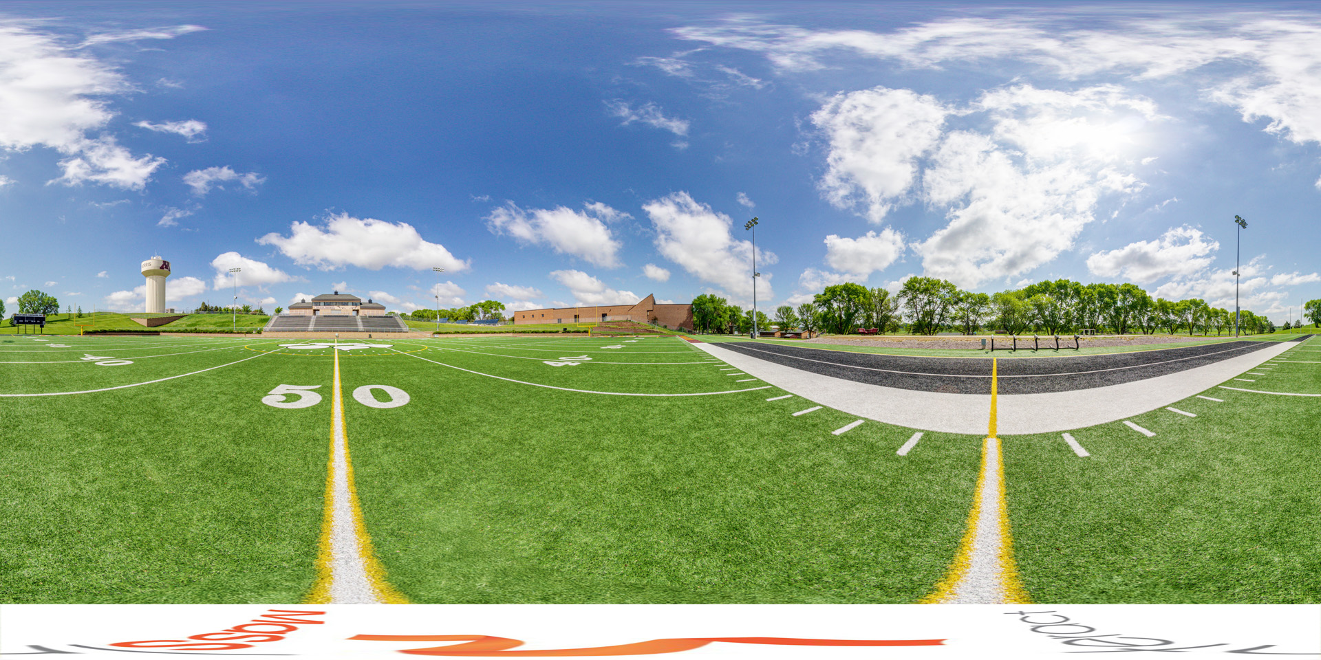 Panoramic view of a football field at the 50-yard line with bleachers and a scoreboard in the background under a sunny sky.