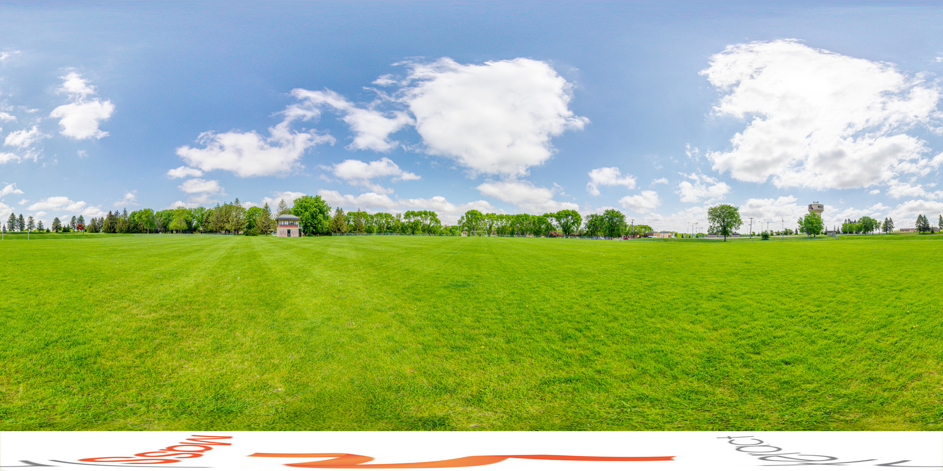 Panoramic view of a wide grassy field under a blue sky with scattered clouds, with trees and buildings visible in the distance at Cougar Field.