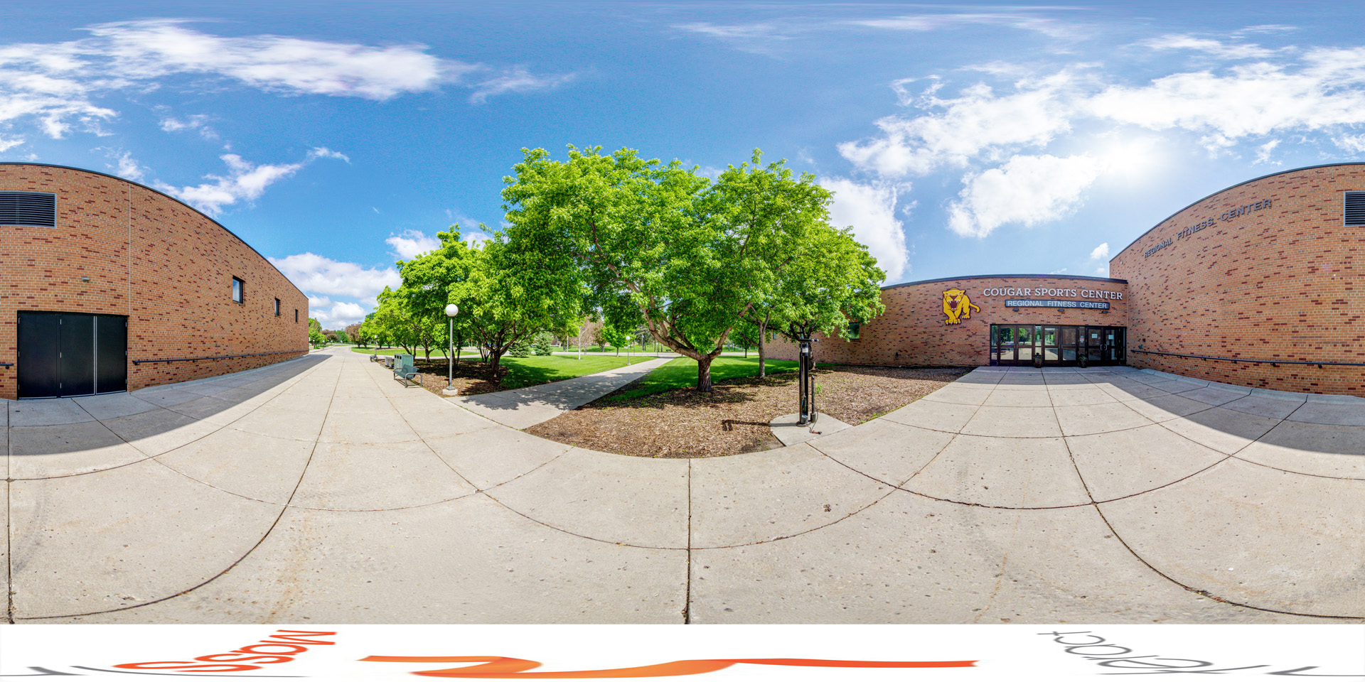 Panoramic view of the entrance to the Cougar Sports Center, featuring a brick building, a pathway, and green trees under a sunny sky.