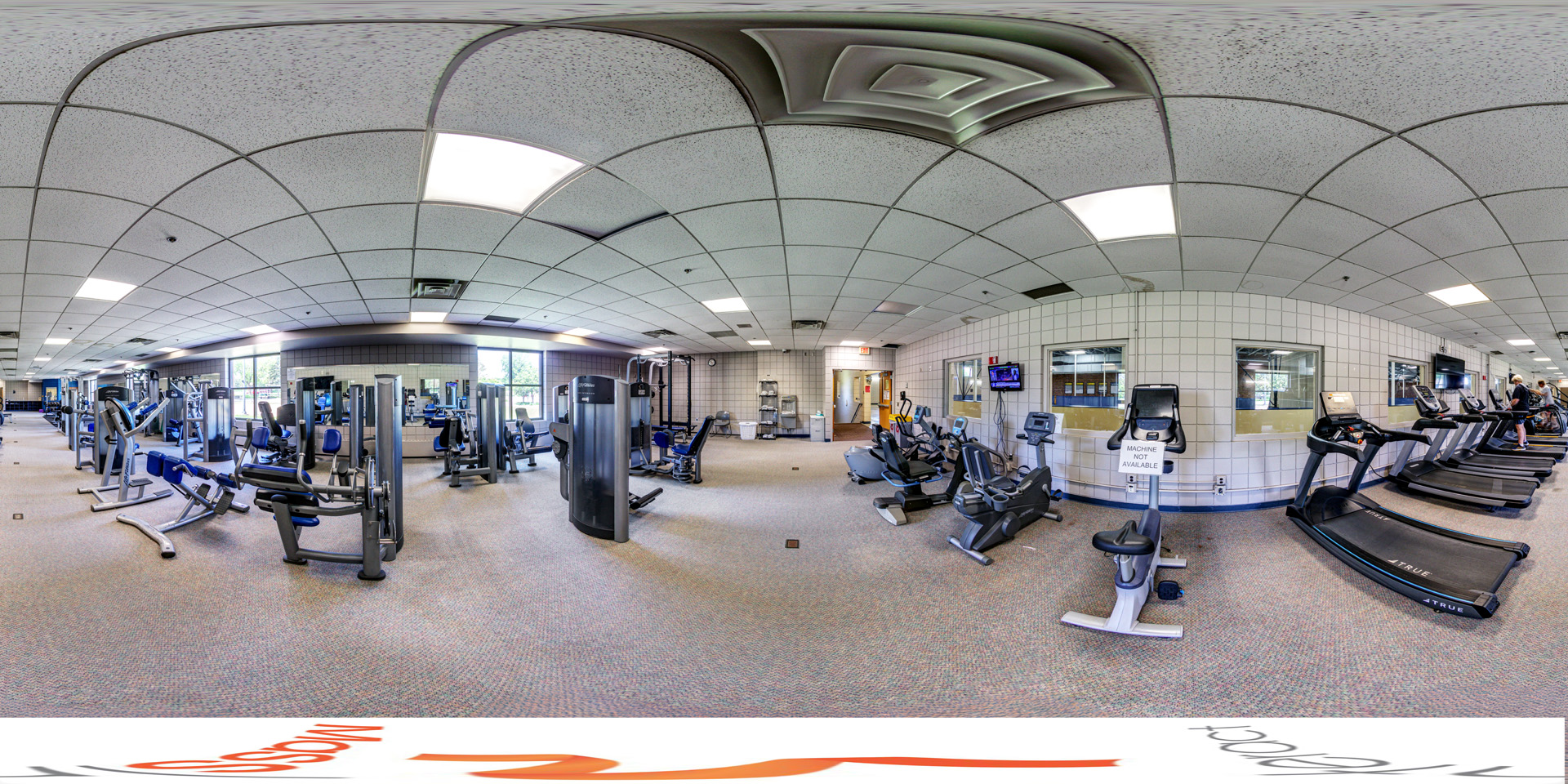 Panoramic view of a fitness room with various exercise machines, including treadmills, stationary bikes, and strength training equipment, with natural light coming through large windows.