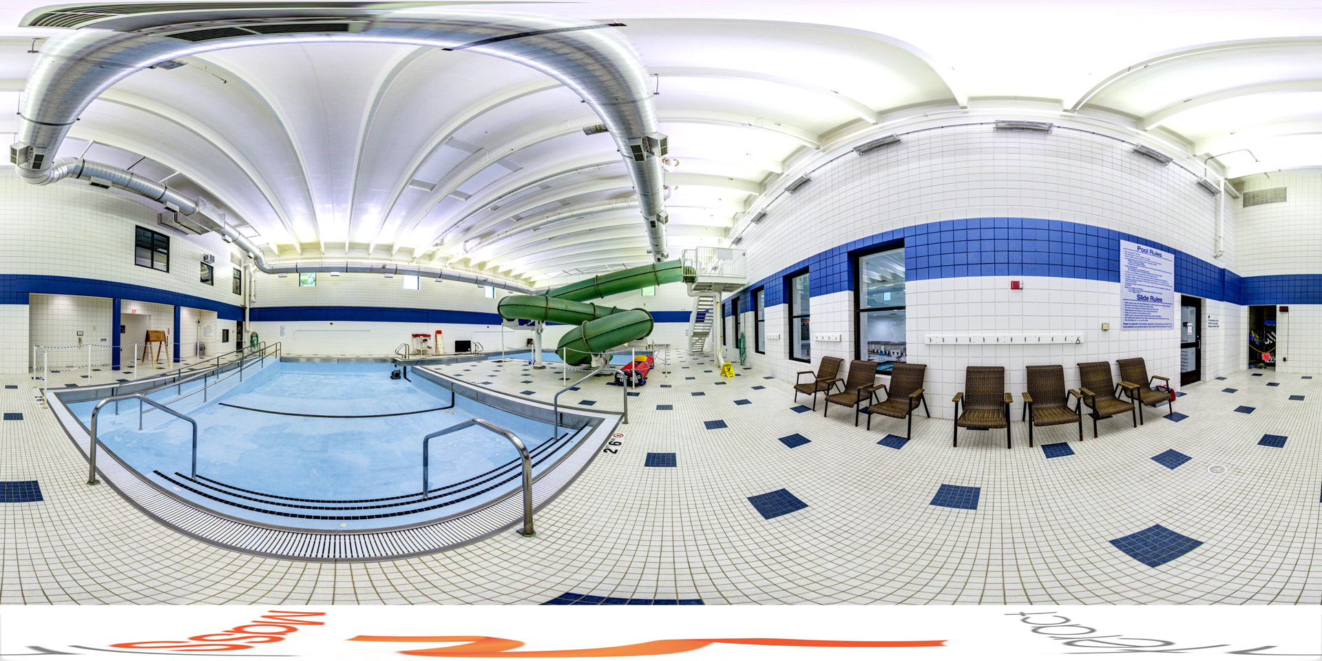 Panoramic view of an indoor recreation pool with white and blue tile walls, a shallow section with handrails, and a large green water slide that curves into the pool. Poolside seating includes several wicker chairs, and safety signage is visible on the walls.