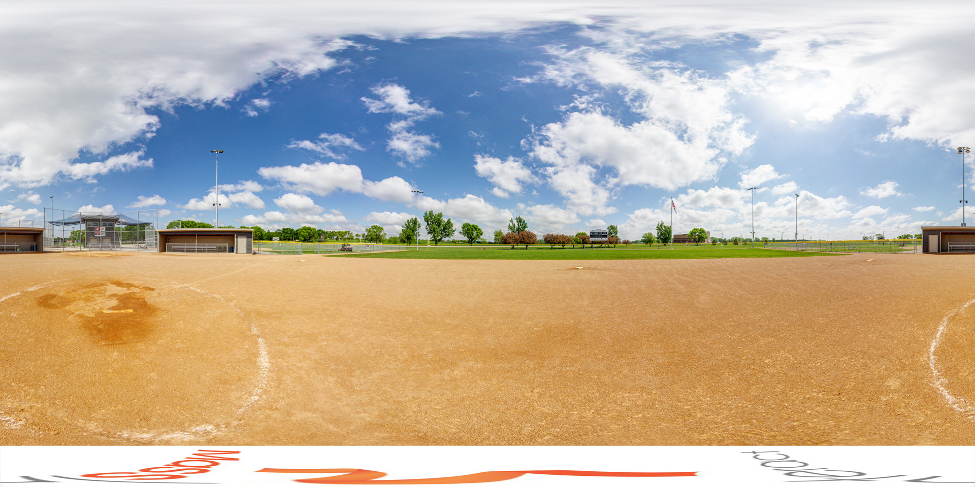 Panoramic view of A softball field with a dirt infield, flanked by two dugouts, and a fenced backstop with bleachers in the distance. The field is under a bright blue sky with scattered clouds. Trees and a scoreboard can be seen in the background.