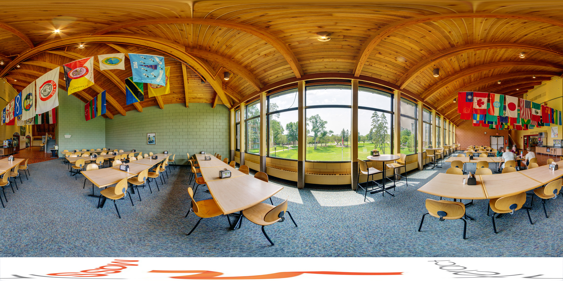 Panoramic view of a brightly lit dining area with long wooden tables and chairs, featuring a large window offering views of a green landscape. Colorful international flags hang from a wooden vaulted ceiling.