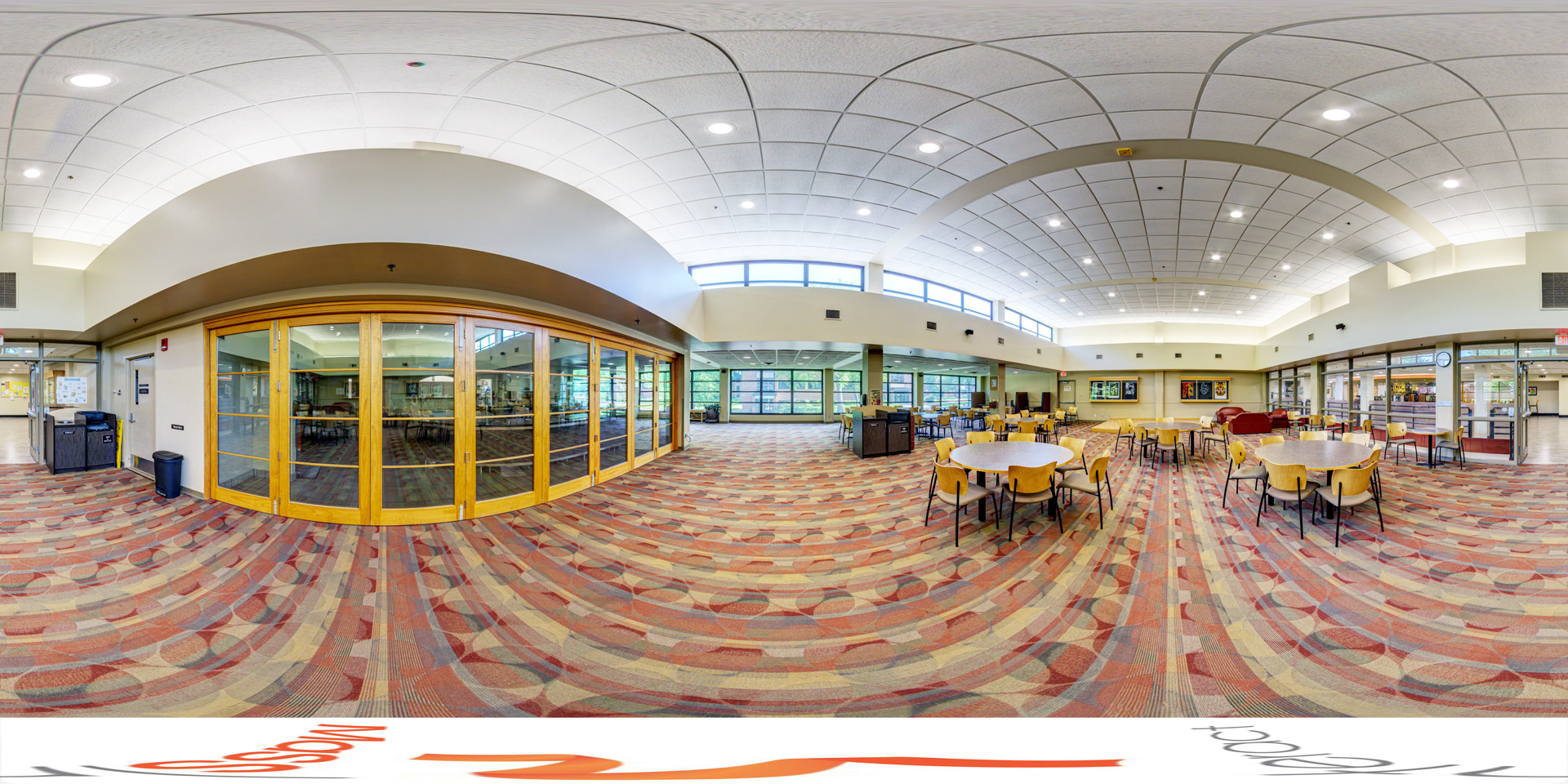 Panoramic view of a Spacious seating area with round tables and chairs, featuring large glass windows and doors that allow natural light. The floor is covered in a patterned carpet, and there are trash and recycling bins near the entrance.
