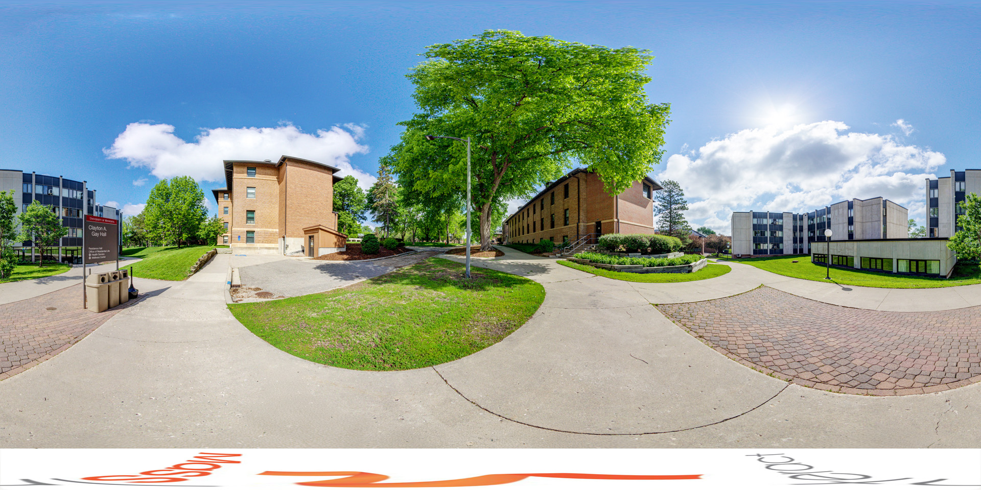 Panoramic view of a Outdoor view of Clayton A. Gay Hall with a pathway leading between brick and modern residential buildings, surrounded by trees and green spaces. A sign for the hall is positioned on the left near trash receptacles.