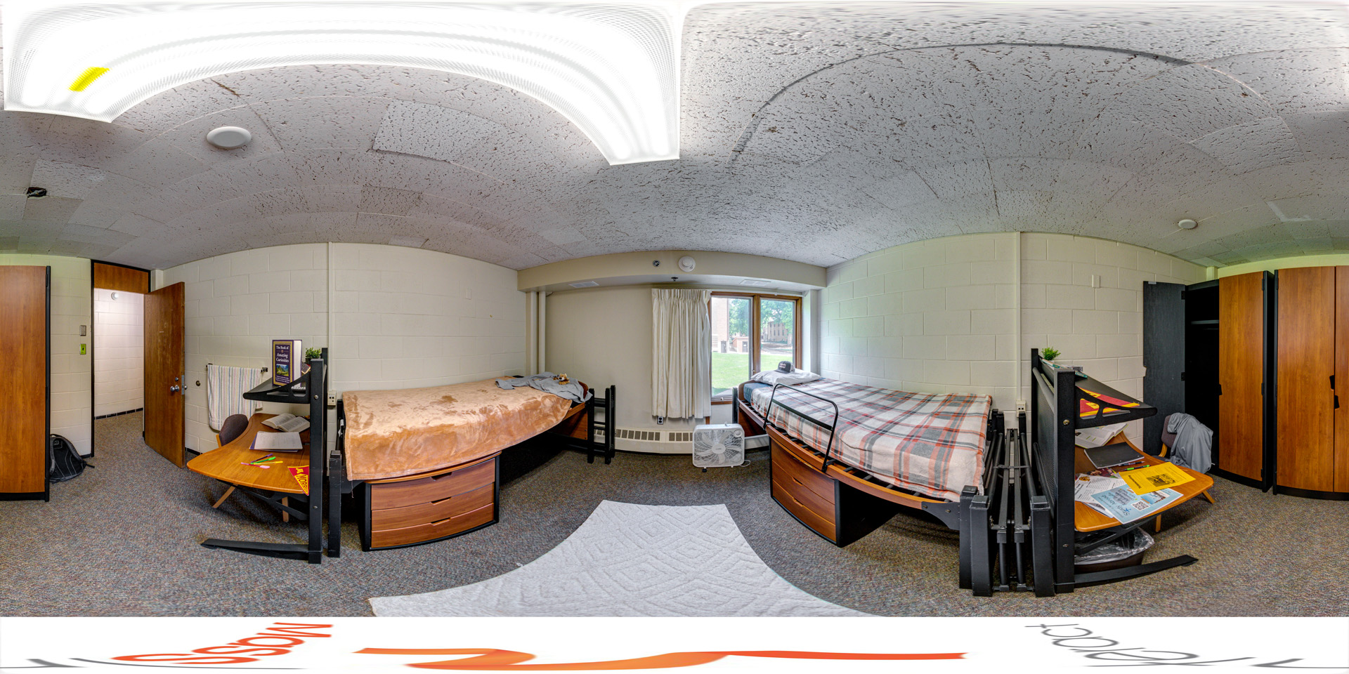 Panoramic view of a double occupancy dorm room with two lofted beds, desks underneath, and wooden wardrobes on either side. The room has a window with a view of trees and a building outside, and personal items are scattered on the desks and beds.