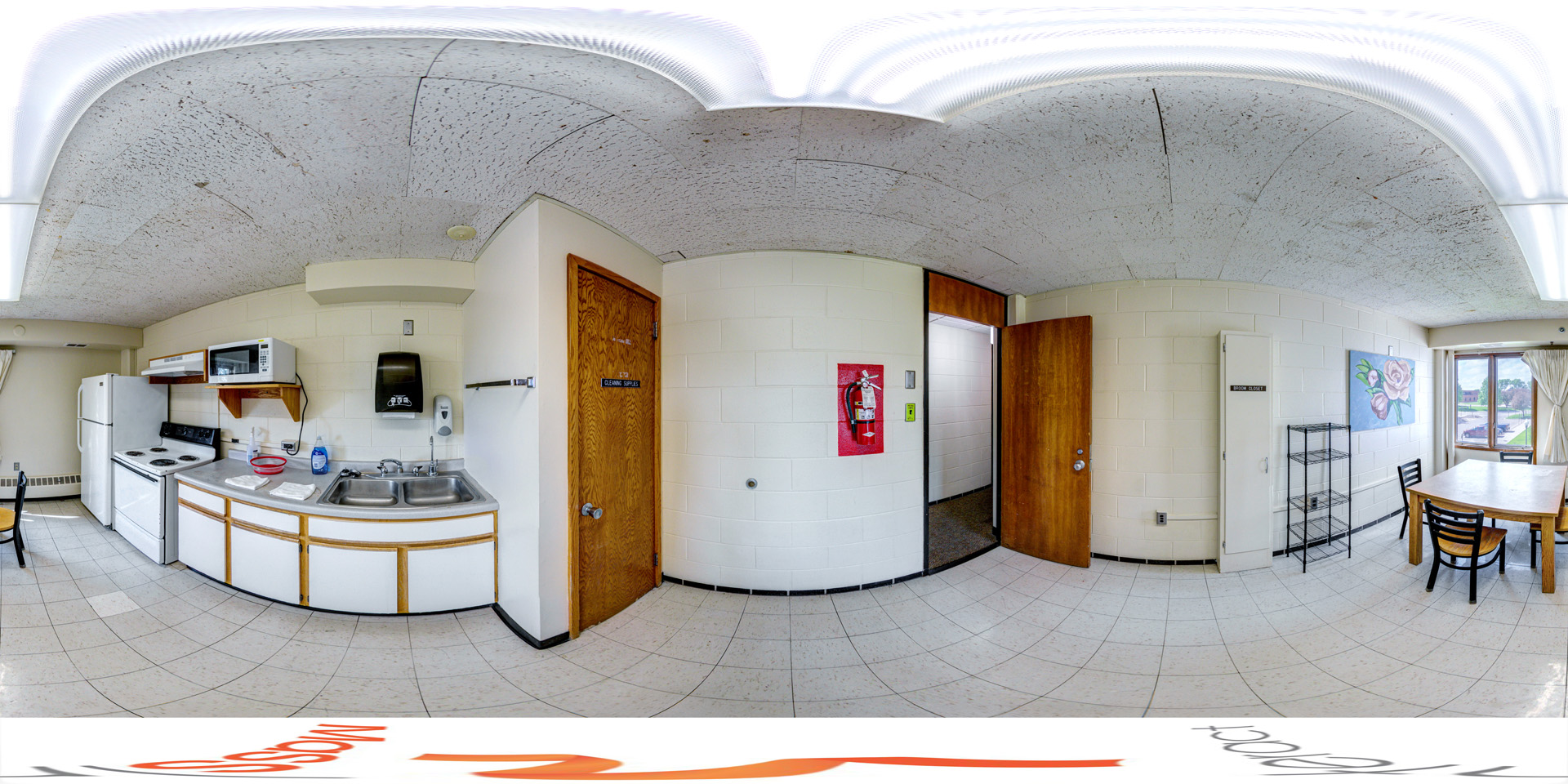 Panoramic view of a shared floor kitchen with a white refrigerator, stove, microwave, and dual sink. There is a wooden dining table with chairs next to a window that offers a view of the outdoors. The kitchen is simple, with tiled floors, basic cabinetry, and a fire extinguisher mounted on the wall.