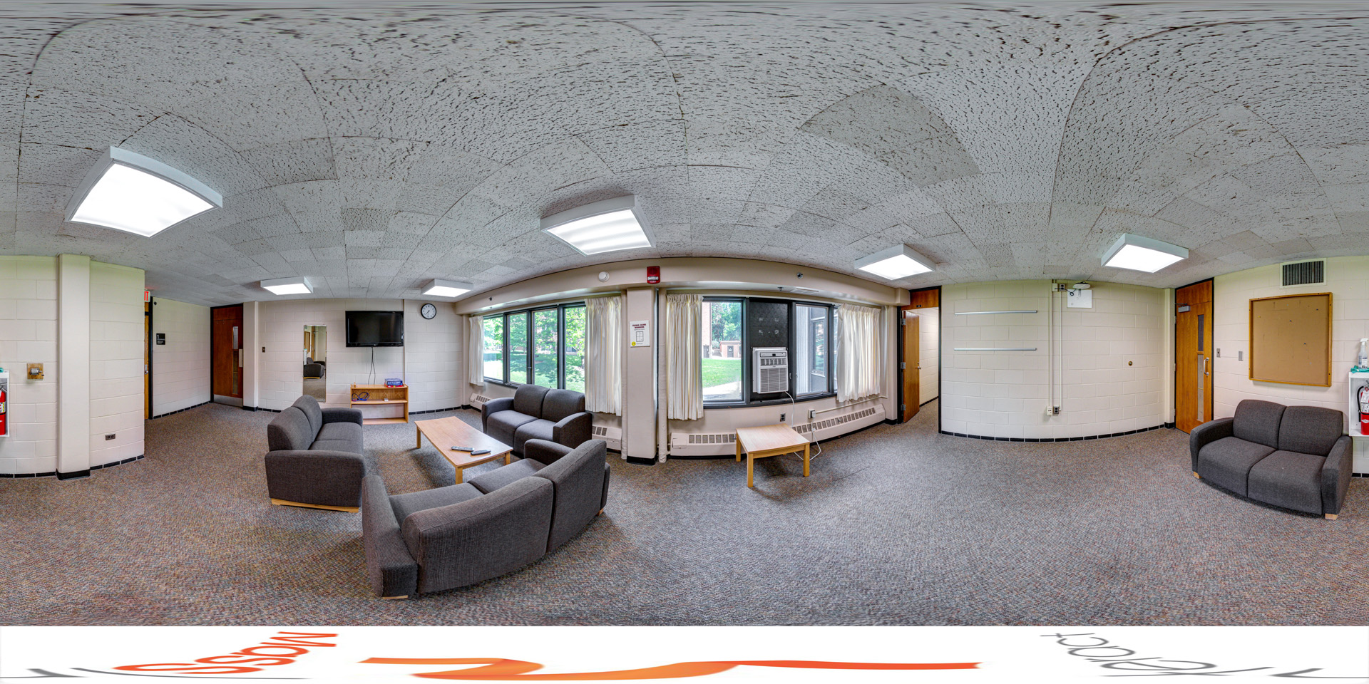 Panoramic view of a cozy floor lounge with gray upholstered couches and a wooden coffee table set in the center. The room is carpeted and features large windows with curtains, offering views of greenery outside. A mounted TV and a clock are positioned on the wall, and there is an air conditioning unit below the windows.