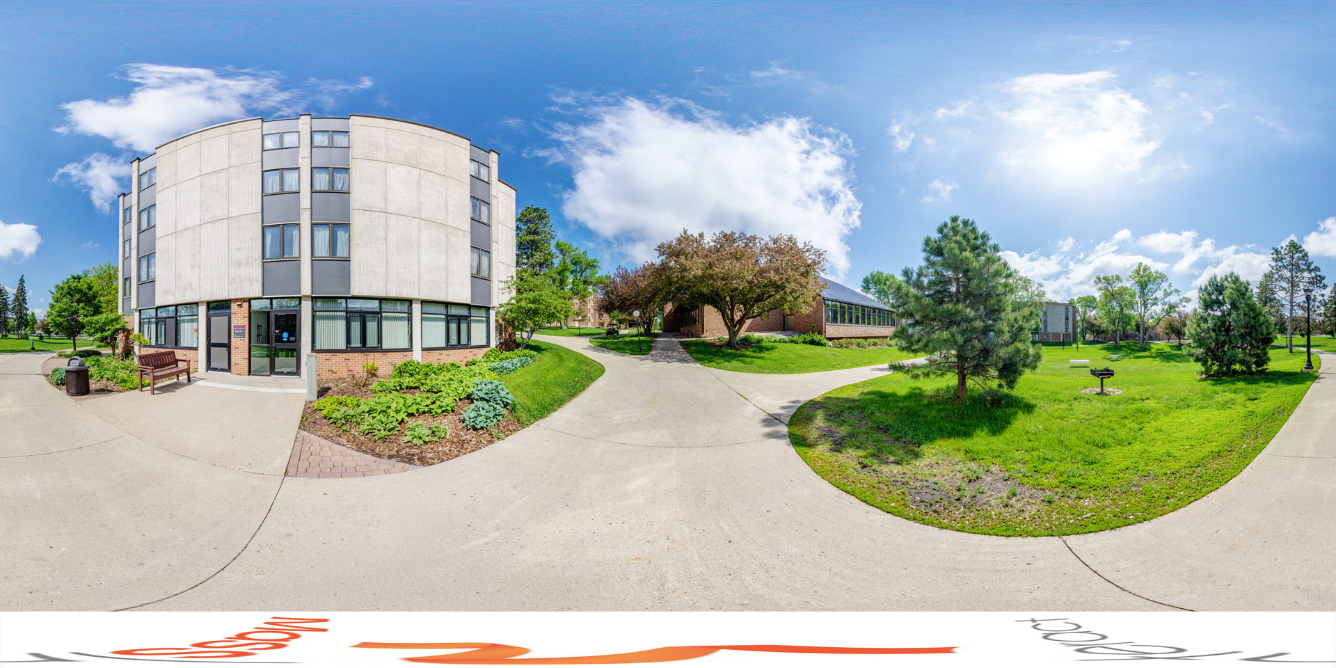 Panoramic view of a sunny day outside a modern building with a mix of concrete and brick. The entrance is surrounded by greenery, with a bench nearby. Walkways lead to the building and other areas, lined with trees and well-kept lawns. A picnic area with grills is visible to the right.