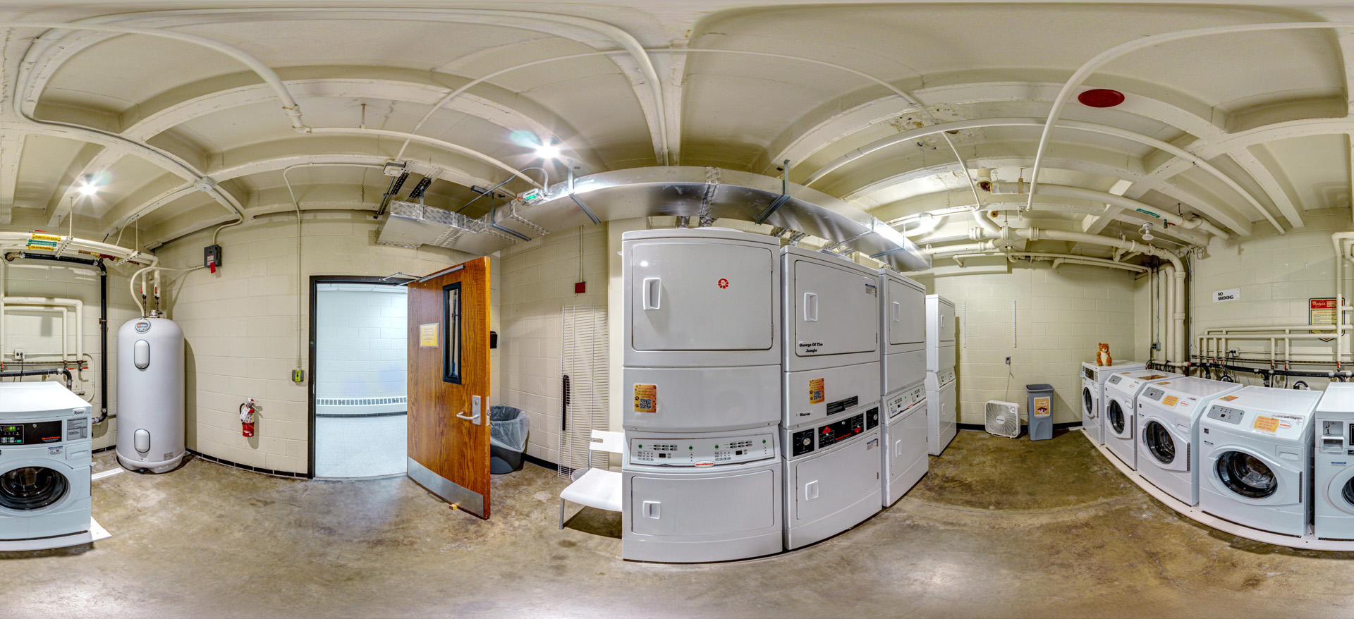 Panoramic view of a residence hall laundry room with stacked dryers, front-loading washing machines, exposed ceiling pipes, concrete floors, and an open doorway leading to the hallway.
