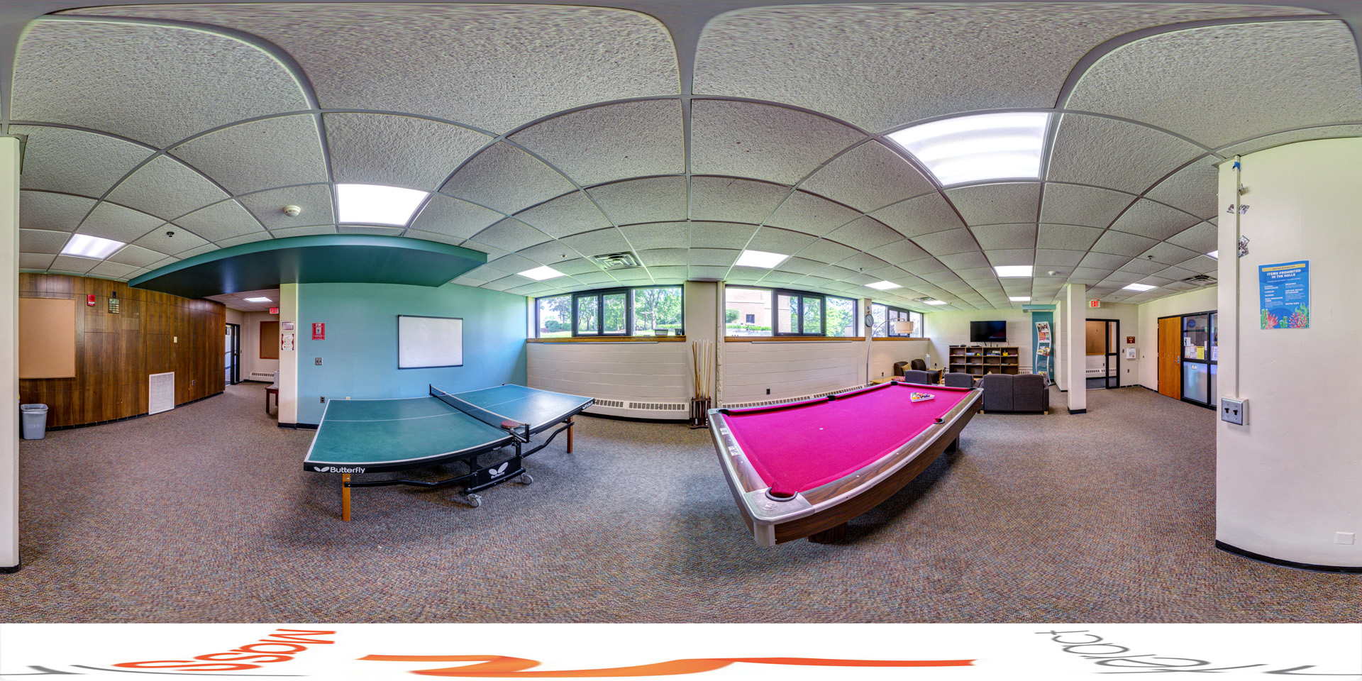 Panoramic view of a recreational lounge area with a pink pool table and a ping-pong table. The room is carpeted and has ceiling tiles with fluorescent lighting. Windows allow natural light in, and there is a small seating area with a TV in the corner. Several doors lead to other parts of the building, and pool cues are stored near the windows.