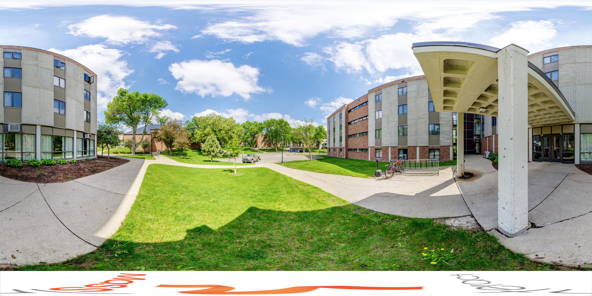 panoramic view of the entrance to David C. Johnson Independence Hall (Indy), a multi-story residential building with modern concrete and brick facades. A bike rack sits near the entrance, and the surrounding area features green lawns, trees, and a network of walkways connecting to nearby buildings. 