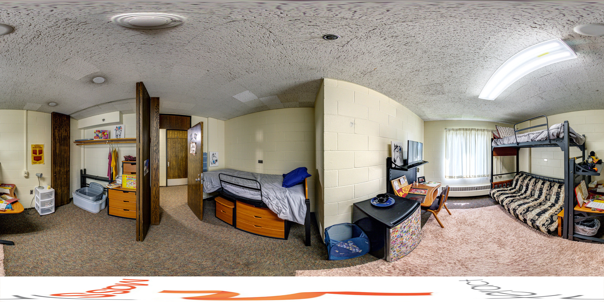 Panoramic view of a cozy double-occupancy dorm room with bunk beds, desks, and shelving. The lower bunk is a futon, and a window brings in natural light. The space is compact yet functional, with a small refrigerator and a shaggy rug for added comfort.