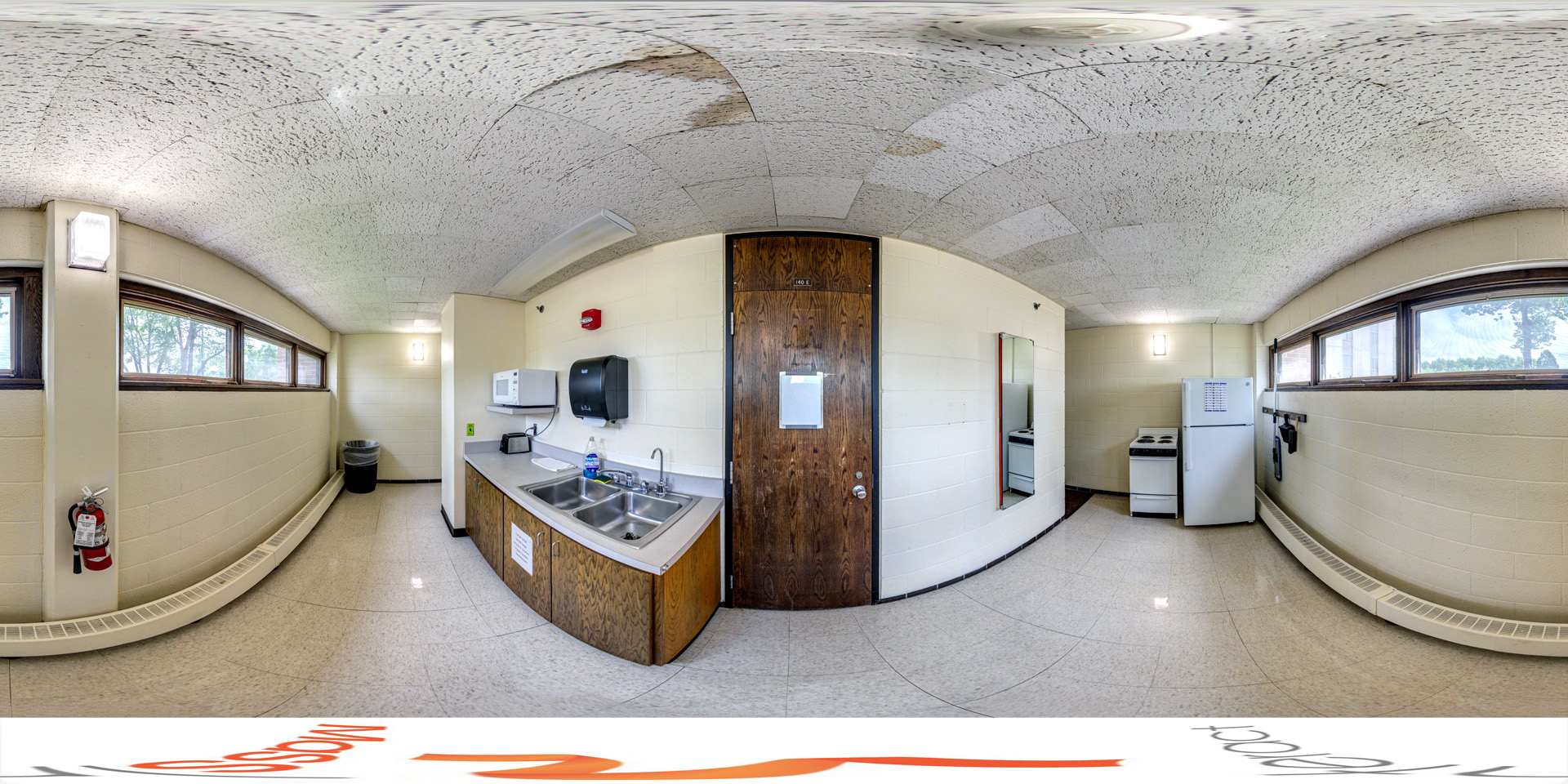 Panoramic view of a small communal kitchen featuring a double sink, a microwave, and a refrigerator. A stove is located in the corner, and the room has upper windows for natural light. The space is simple and functional, with essential appliances for shared use.