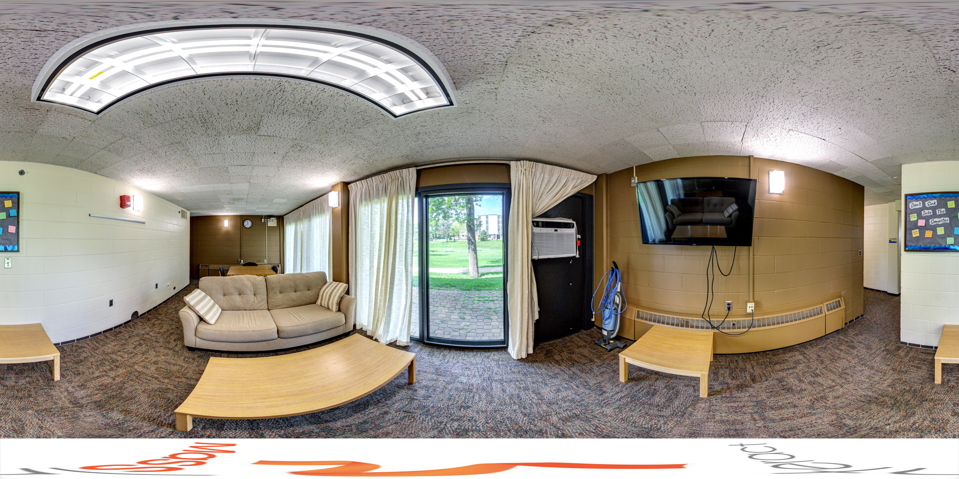 Panoramic view of a cozy lounge area with a beige sofa, a flat-screen TV mounted on the wall, and large windows offering a view of the outdoors. The room features simple furniture, including a coffee table, and a bulletin board for announcements. It has carpeted flooring and an air conditioning unit near the window.
