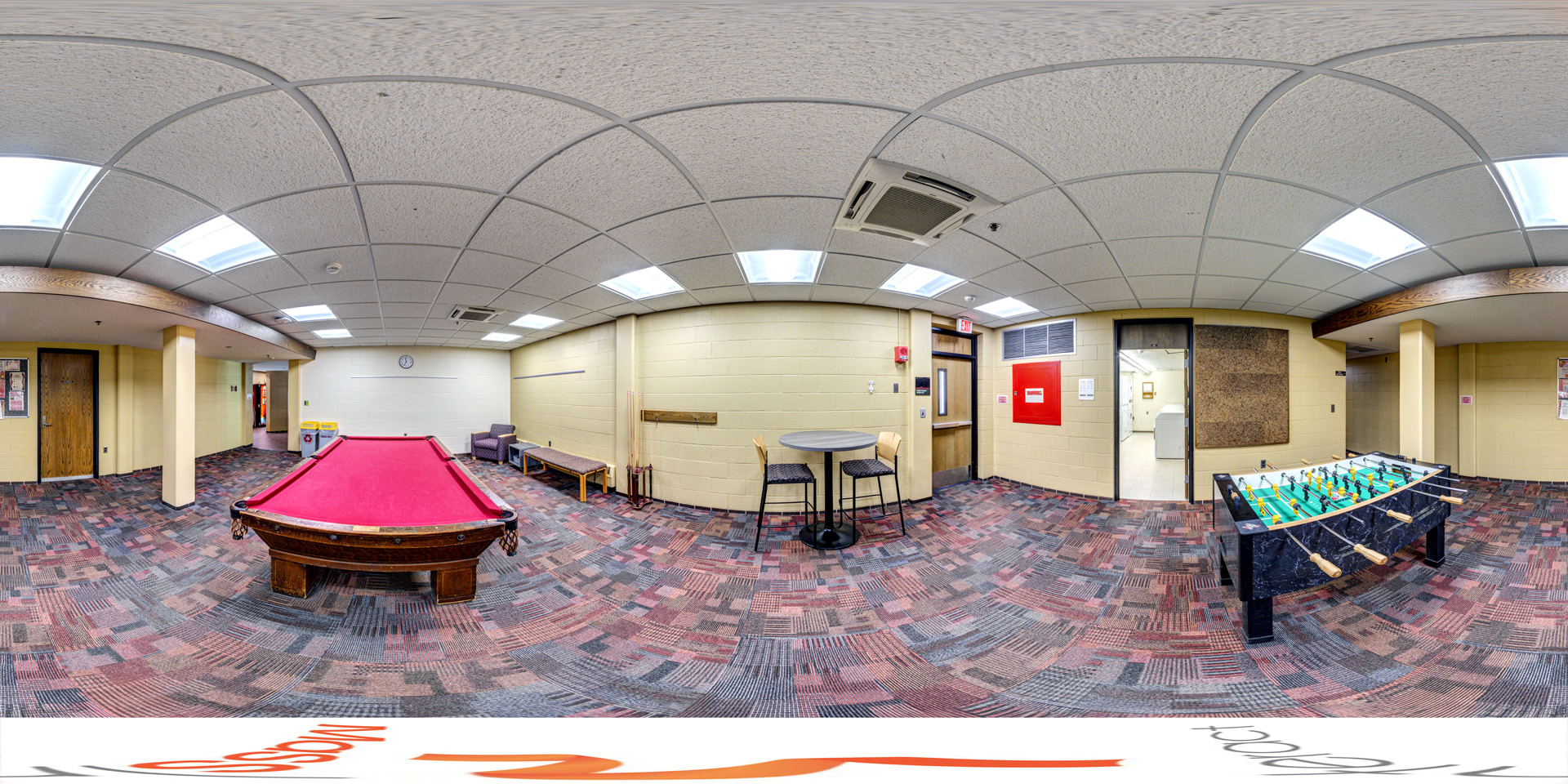 Panoramic view of a recreation room with a red-felt pool table at the center and a foosball table on the side. The room features patterned carpet flooring, a small round table with two chairs, and a laundry room visible through an open doorway. The space is brightly lit with a neutral color palette and overhead lighting.