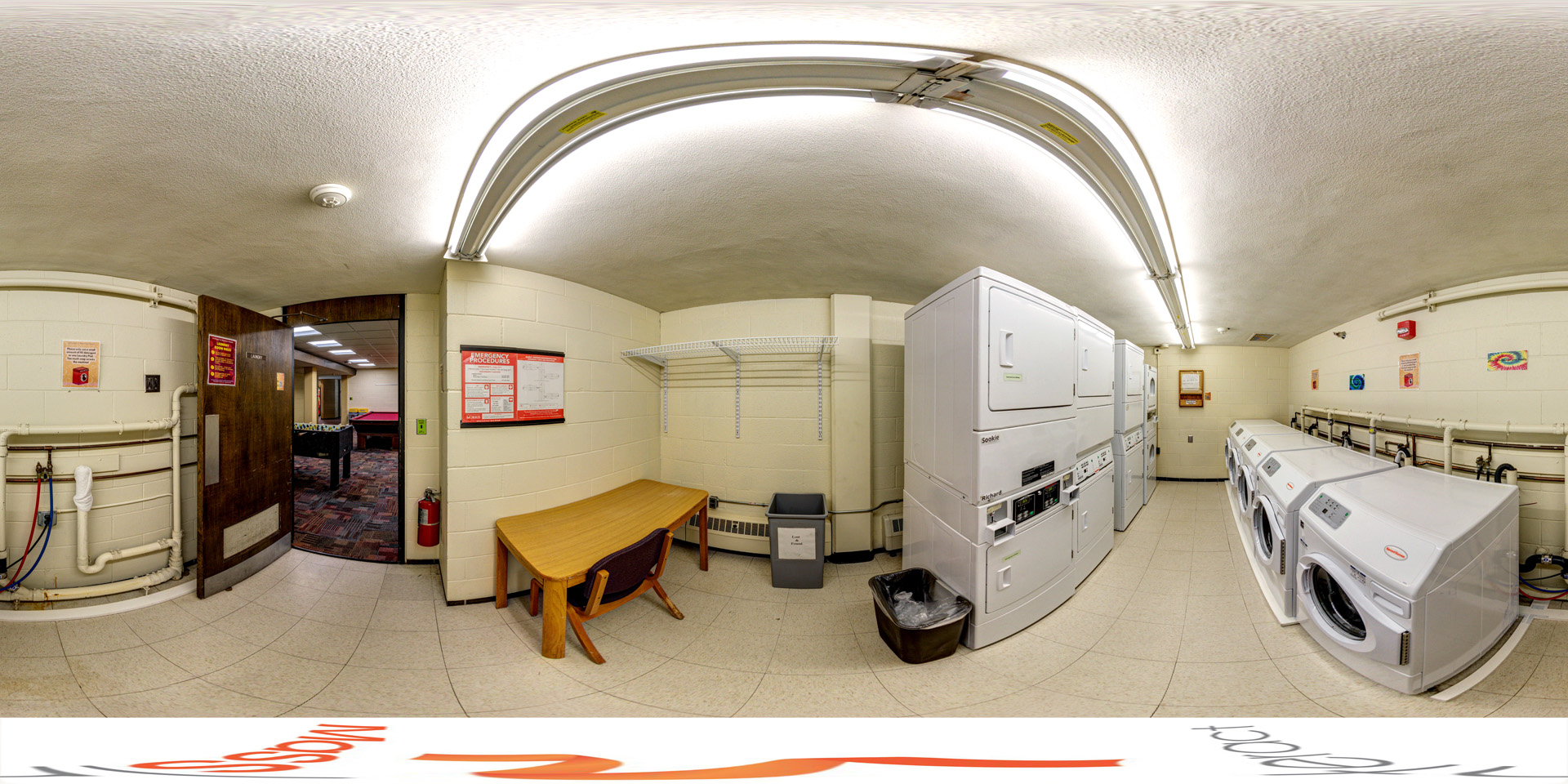 A panoramic view of a laundry room with white cinder block walls, tiled floors, and fluorescent lighting. Multiple front-loading washers and dryers are lined along the right wall. A wooden table with a chair and a waste bin are on the left.