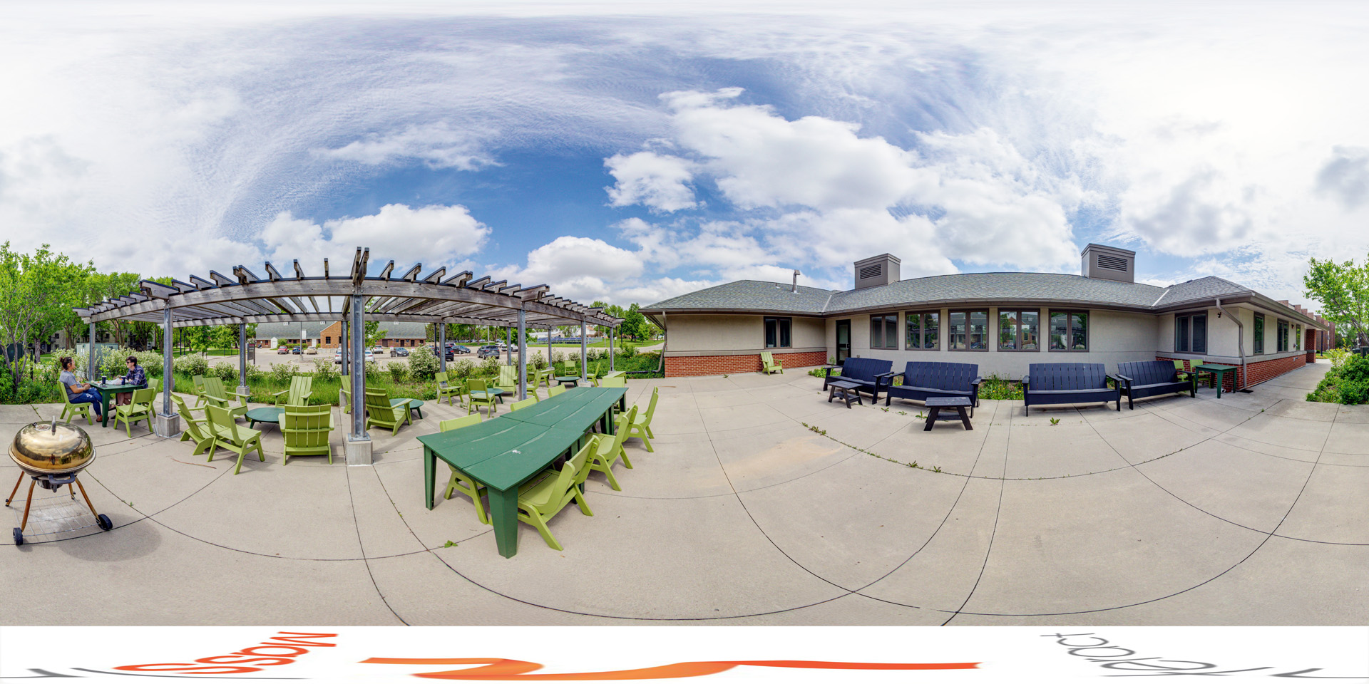Panoramic view of an outdoor seating area featuring green Adirondack chairs arranged around tables under a wooden pergola. The space is part of the Green Prairie Community, with a building to the right featuring large windows. A grill is also visible in the foreground, and the surrounding landscape includes lush greenery under a partly cloudy sky.