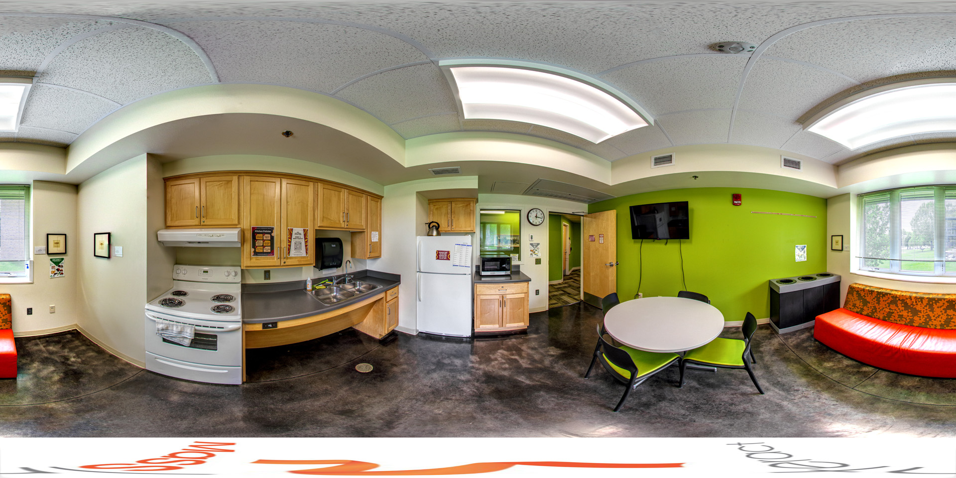 Panoramic view of a small kitchen and lounge area featuring a stove, refrigerator, and sink along with light wood cabinets. The walls are painted in shades of green, and the lounge includes a round dining table with green chairs, a bright orange cushioned bench, and a wall-mounted TV.