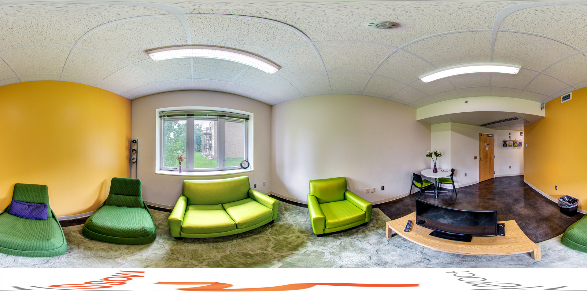 Panoramic view of a cozy lounge with modern green seating, a couch, armchair, and cushioned chairs on a soft rug. Bright yellow and white walls, a large window for natural light, and a flat-screen TV on a small table create an inviting atmosphere. A round dining table with chairs sits near the door.